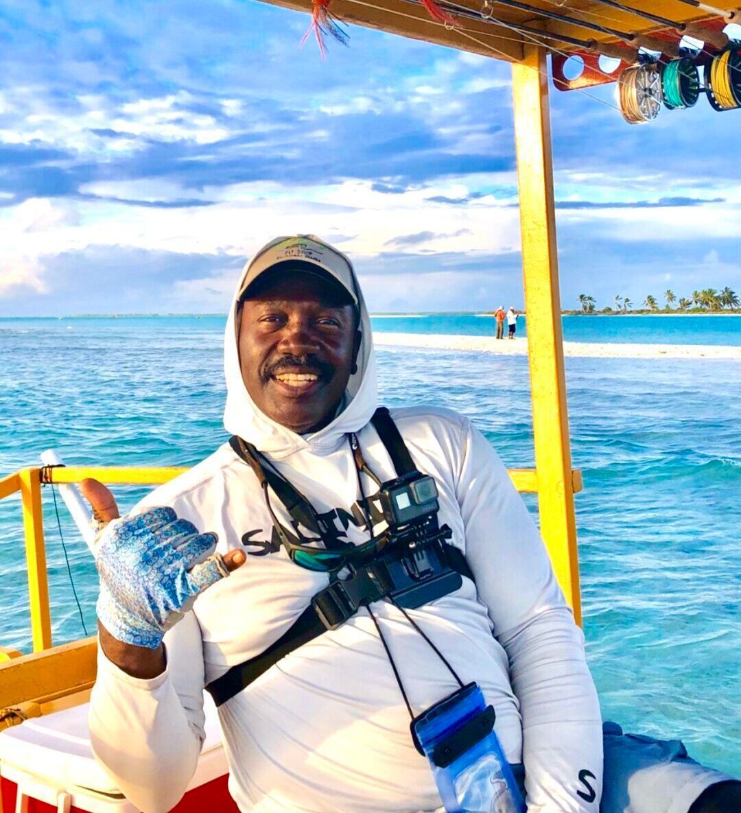 A person wearing a sun hood and fishing gloves smiles and gives a shaka sign on a boat in a bright, tropical setting.
