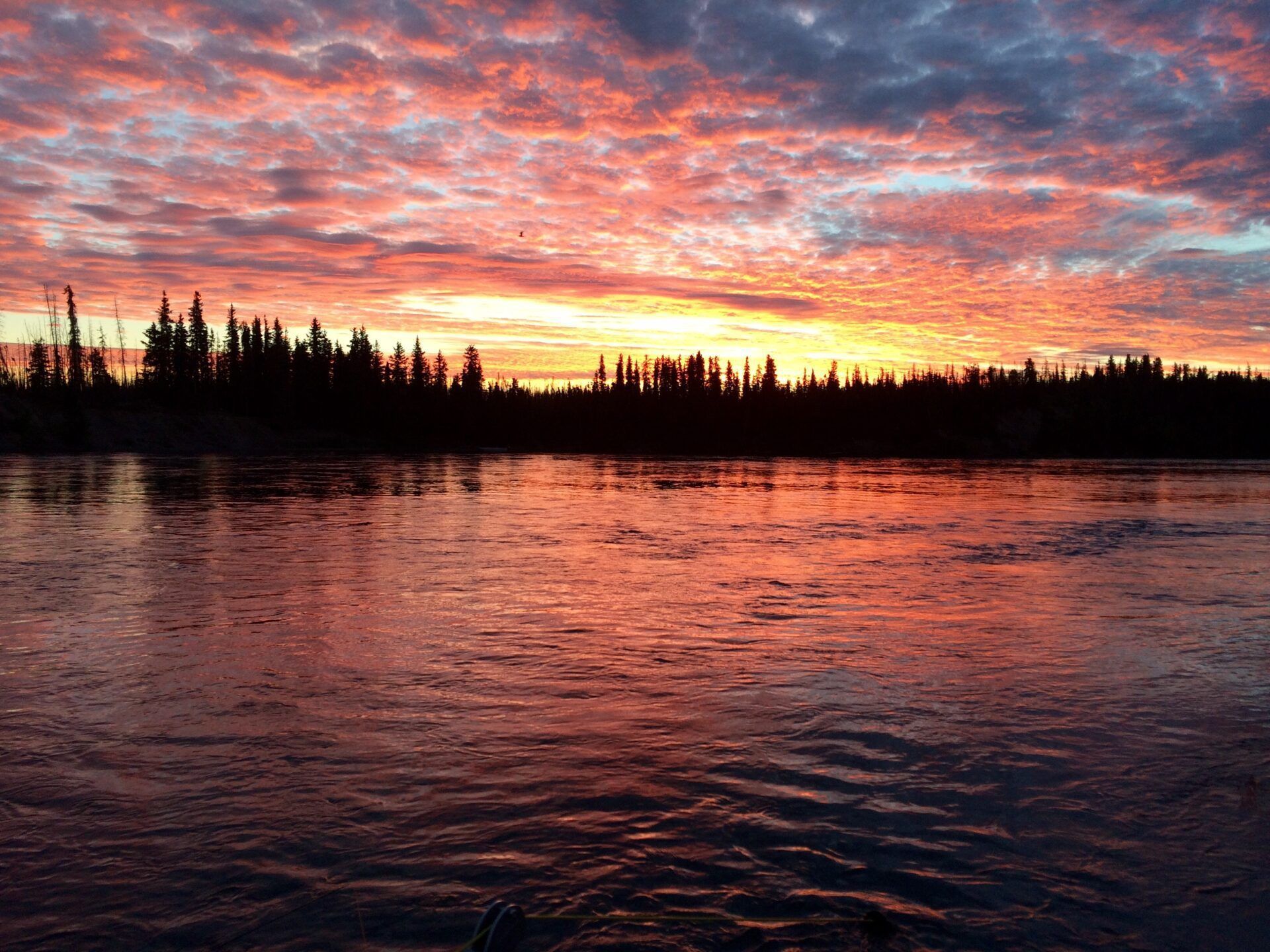 A vibrant orange and purple sunset over a calm river with a silhouetted forest line in the background.