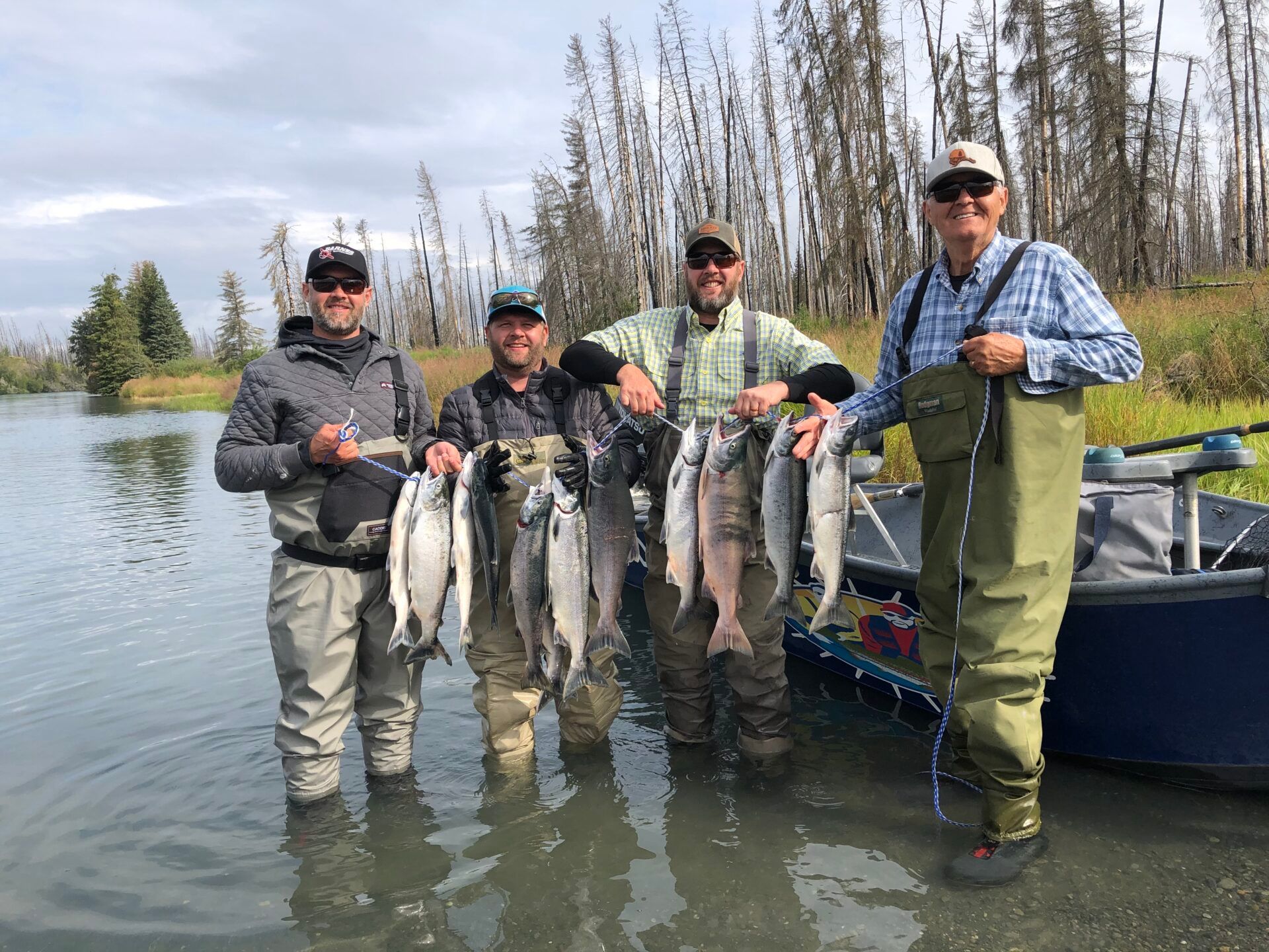 Four people in fishing waders stand in shallow water beside a boat, proudly holding up their catch of several fish.