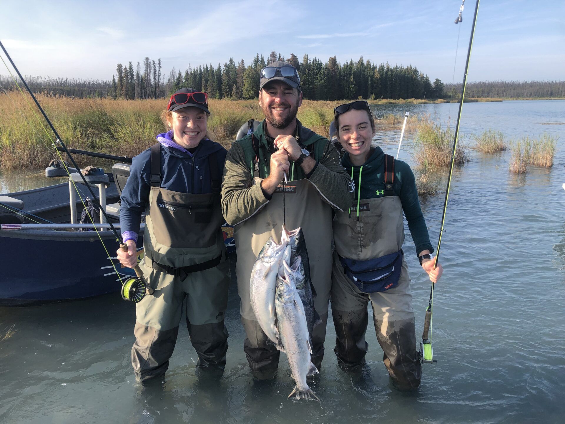 Three people in fishing gear stand in a lake holding two caught fish, with a boat and treeline in the background.
