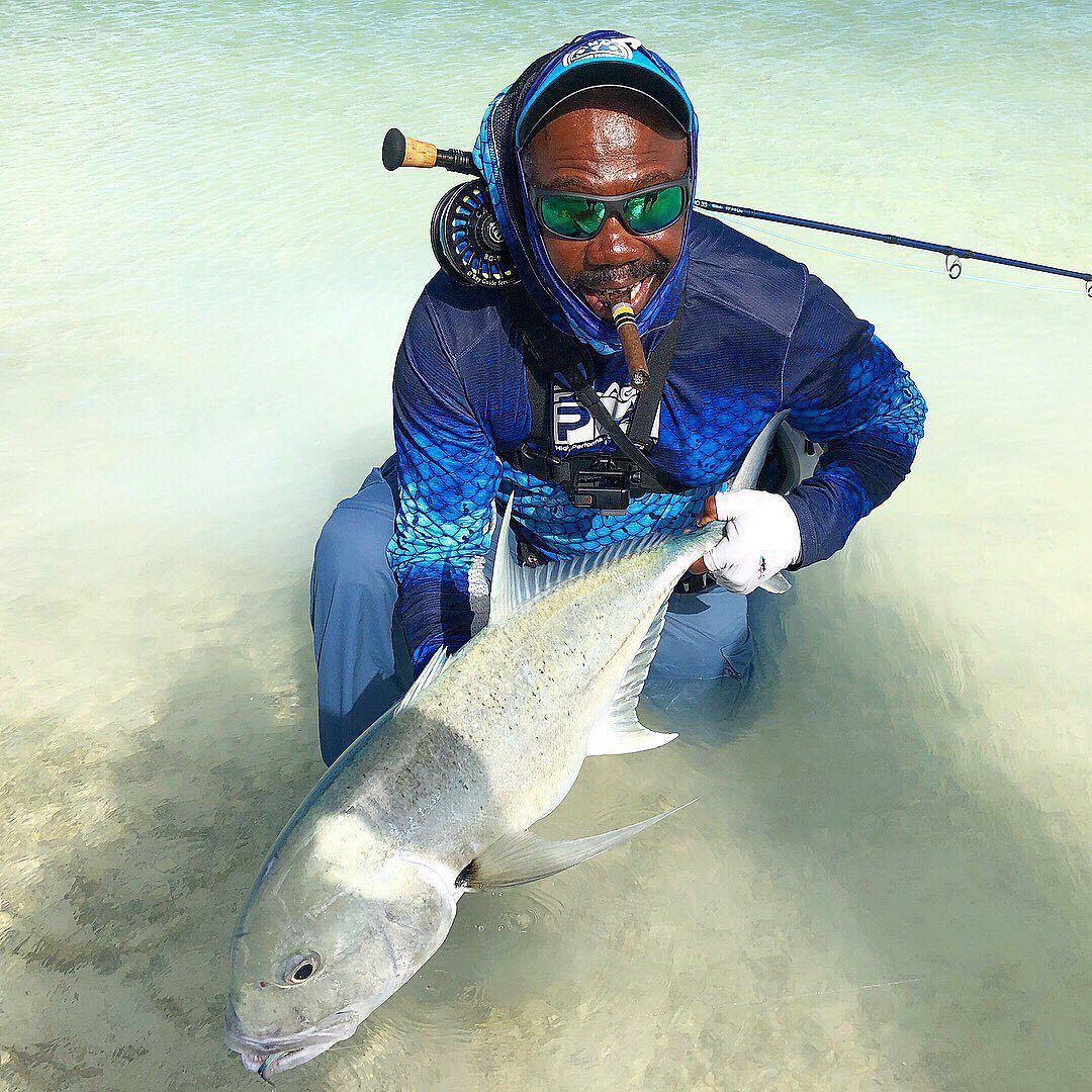 A person wearing fishing gear and sunglasses holding a large fish while standing in clear, shallow water.