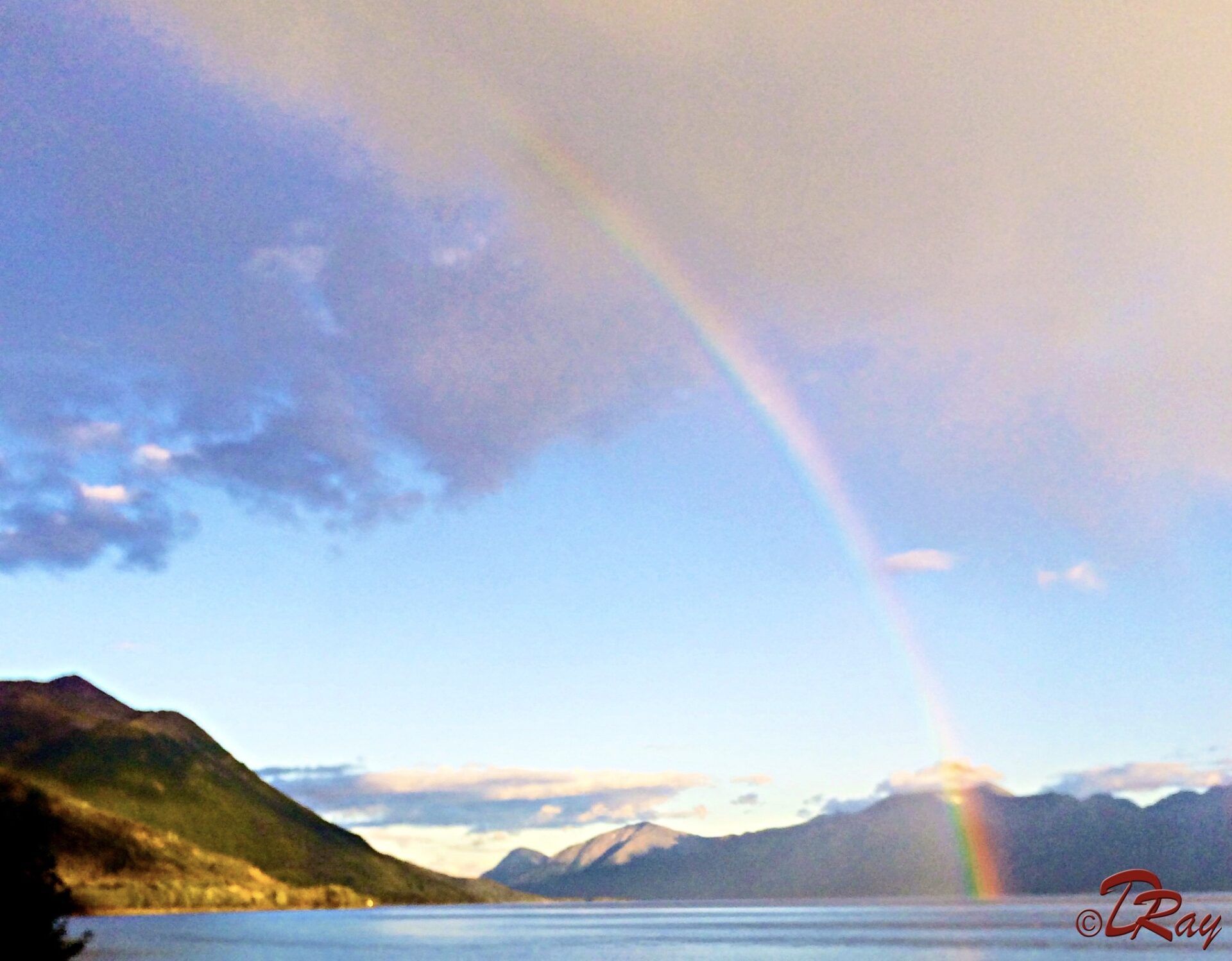 A bright rainbow arches over a calm, expansive lake set against a backdrop of distant mountains under a cloudy sky.