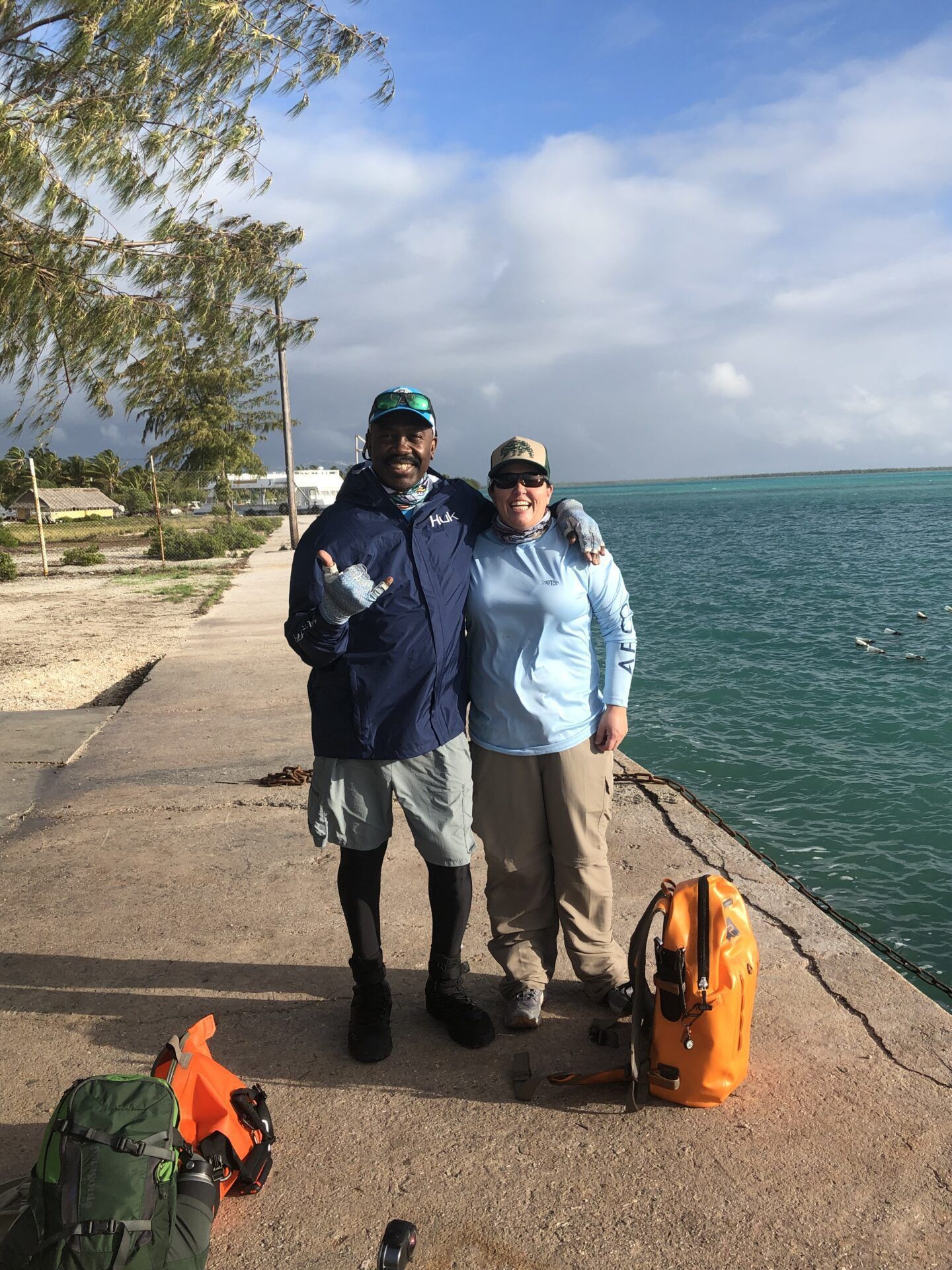 Two people standing on a concrete pier next to the ocean, wearing outdoor gear and hats, with backpacks nearby.