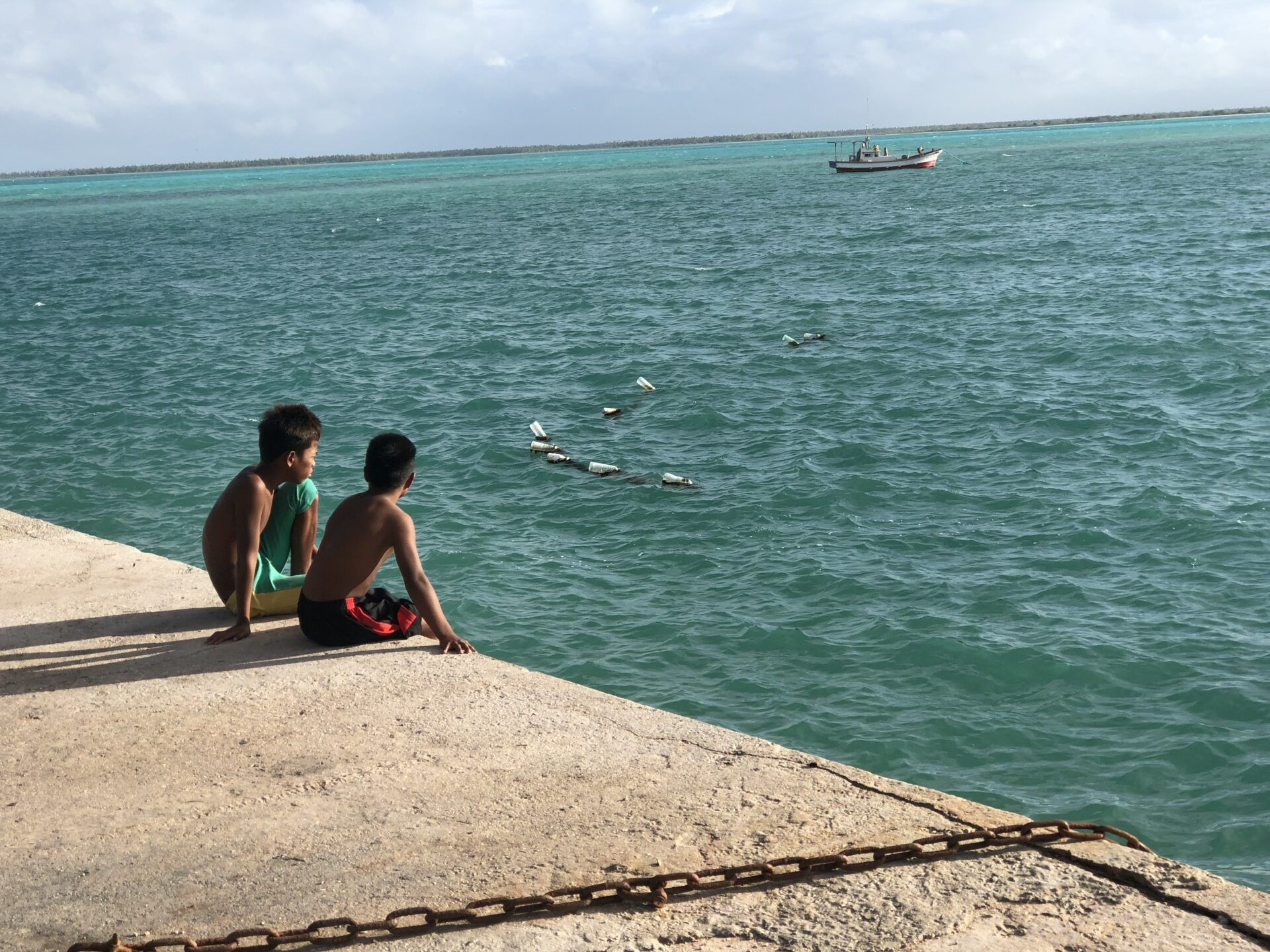 Two people sit on a concrete pier, looking out over turquoise water at a distant boat and small floating objects.