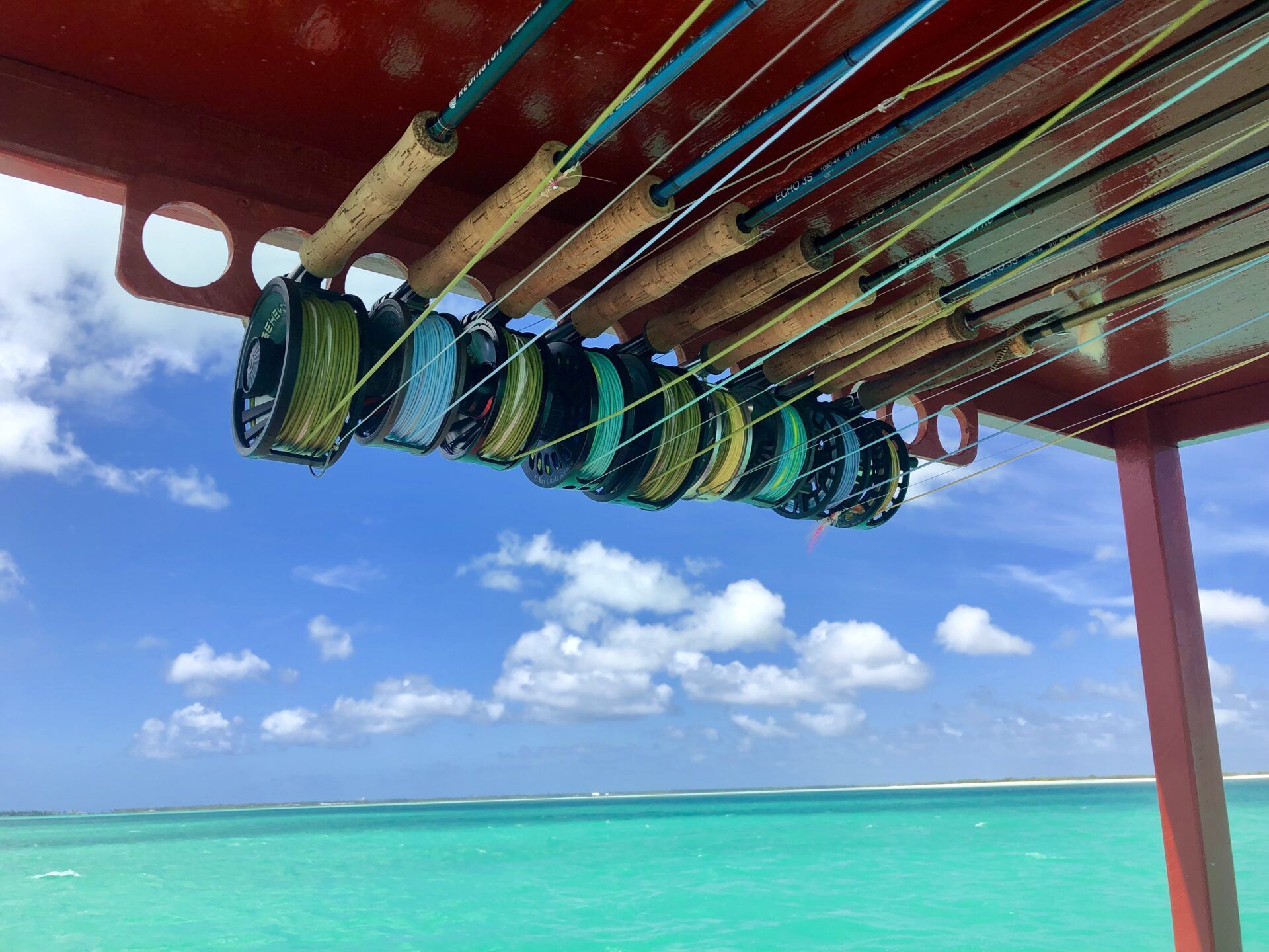 Several fly fishing rods with reels stored on a rack above a boat, overlooking a bright turquoise tropical sea.