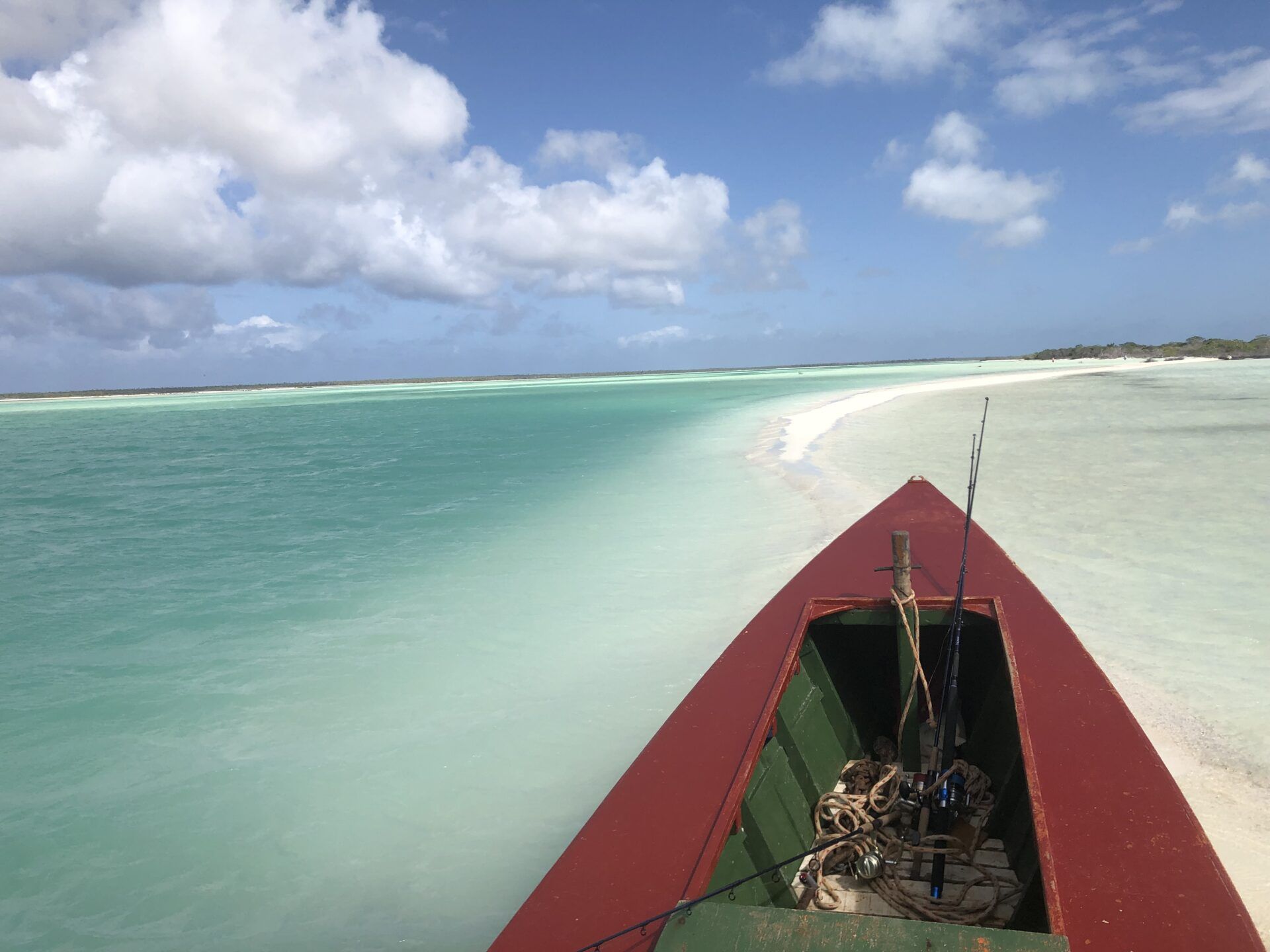 A red boat bow pointing toward a white sandbar in shallow, turquoise water under a blue sky with scattered clouds.