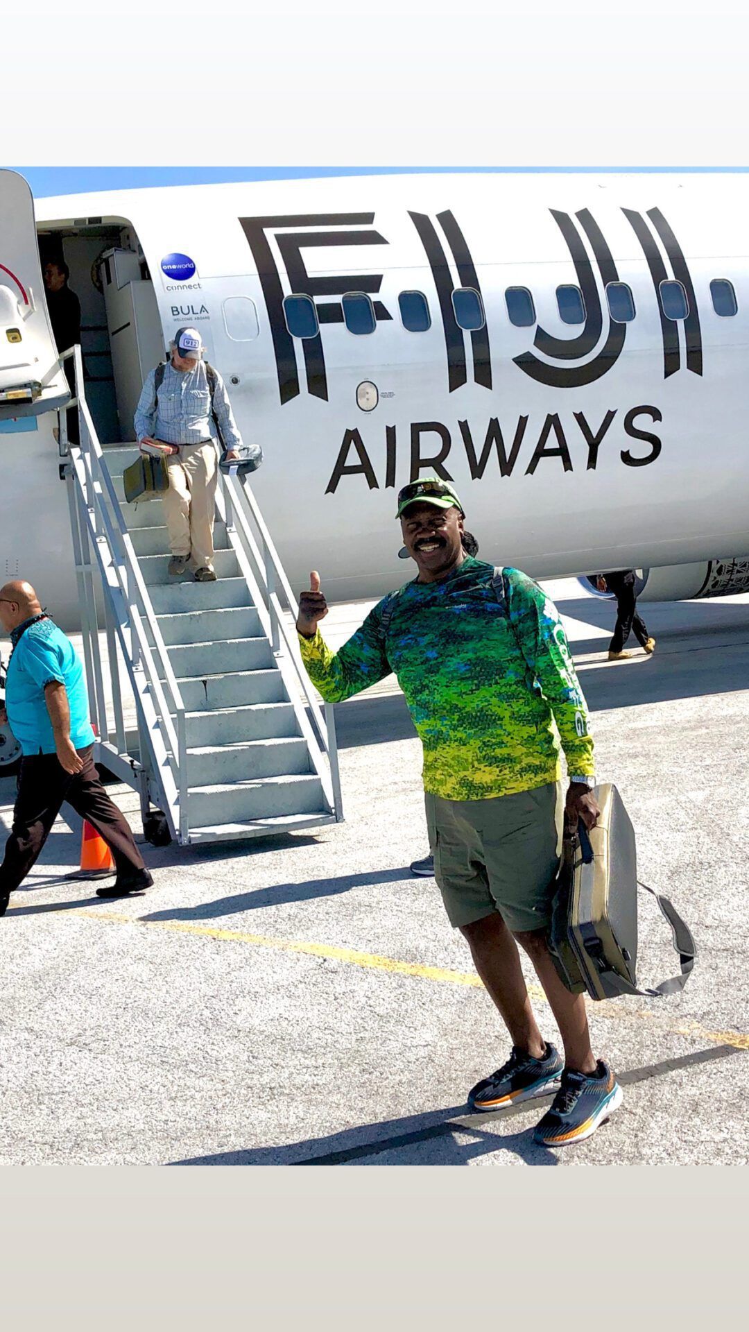 A person wearing a patterned green top gives a thumbs up on an airport tarmac in front of a Fiji Airways plane.