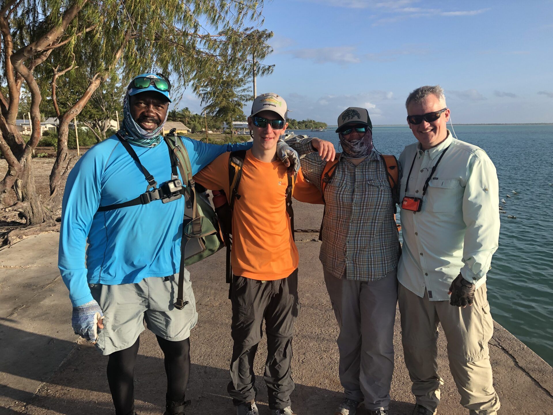 Four people stand side-by-side on a concrete path by the water, wearing casual outdoor gear and smiling.