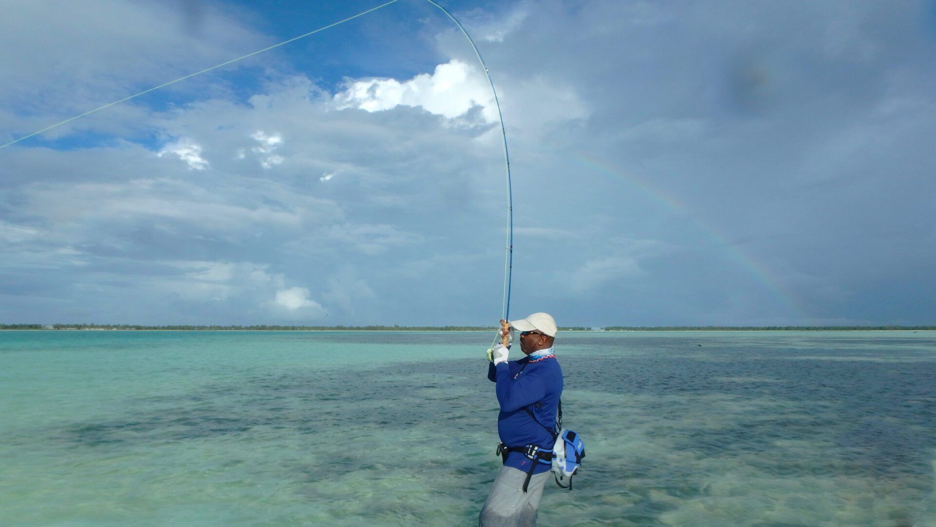 A person in a blue shirt fly-fishing in shallow turquoise water under a cloudy sky.