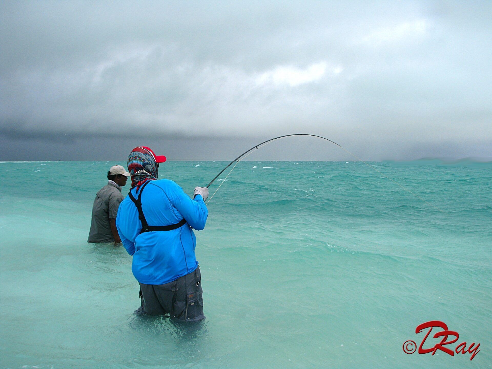 Two people in a shallow, turquoise ocean, with one person casting a fishing line under a dark, cloudy sky.