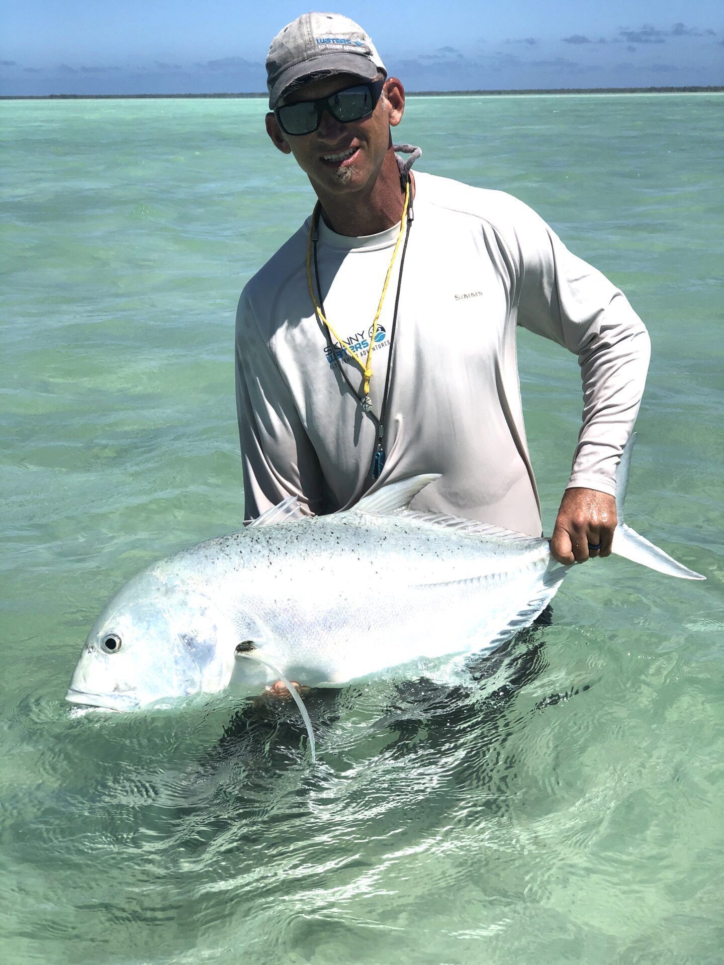A person in a sun shirt and cap holding a large, silver fish while standing in clear, shallow tropical water.