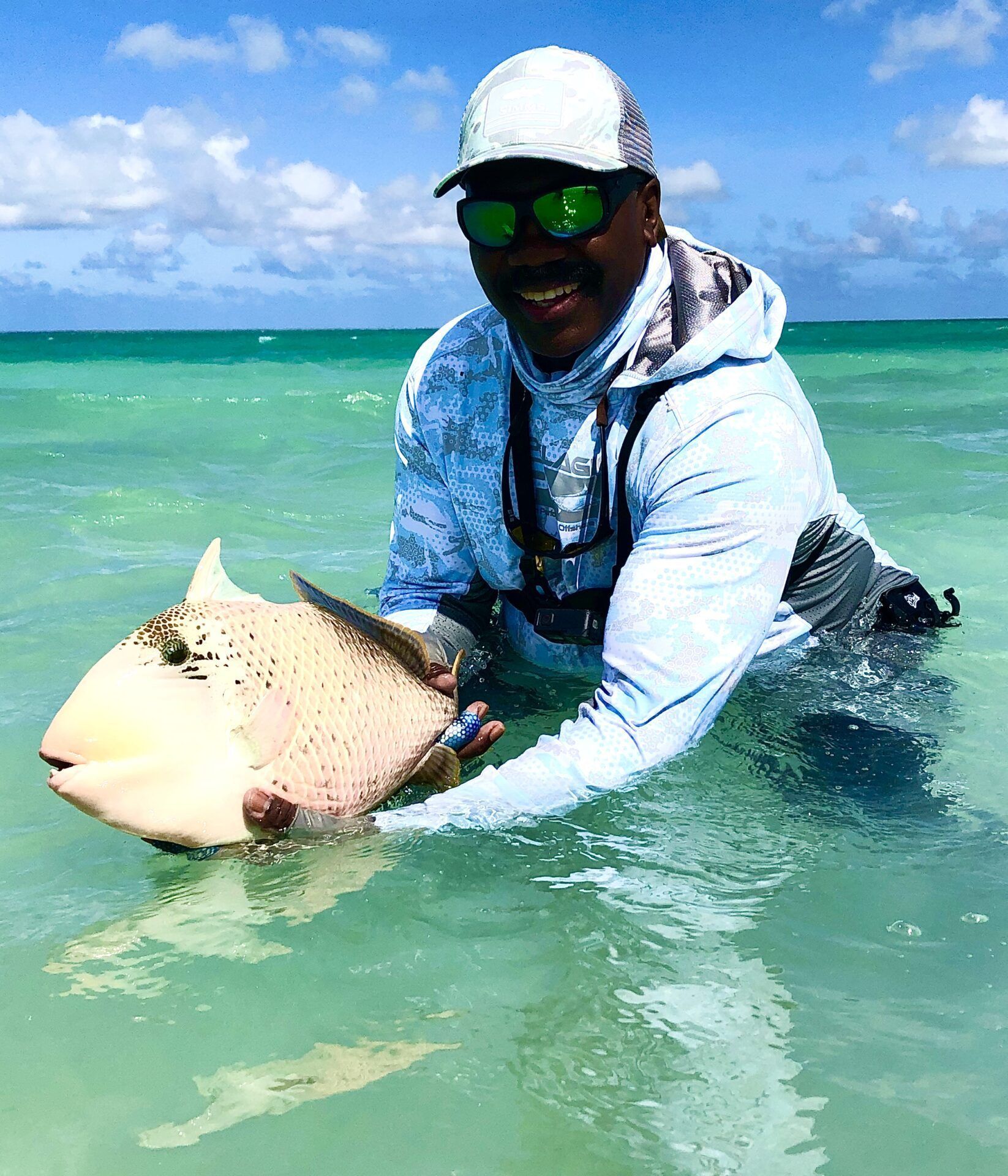 A smiling person wearing a cap and sunglasses holds a large, pale triggerfish in clear, shallow ocean waters.
