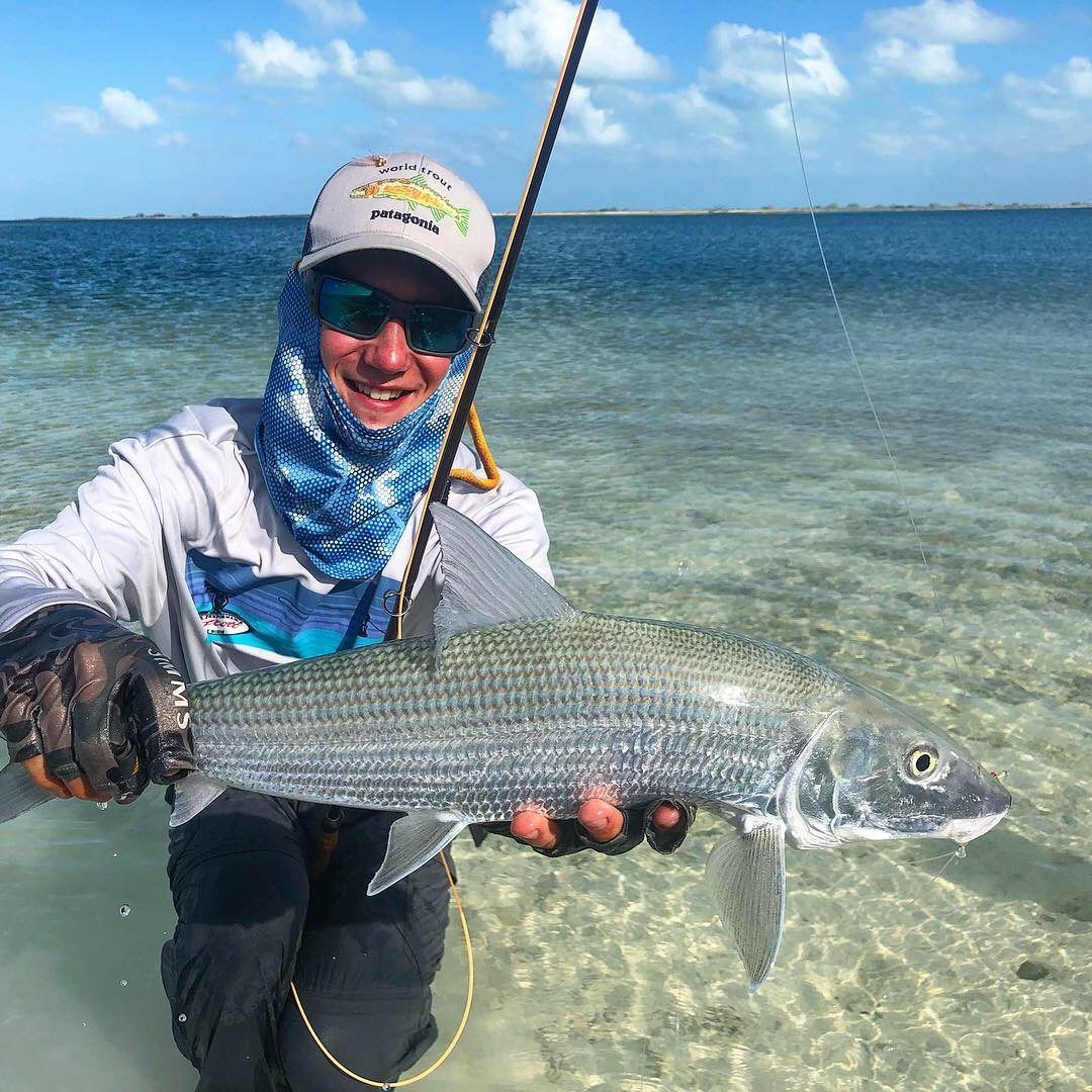 A person wearing sun protection gear kneels in shallow, clear water while holding a silver bonefish.