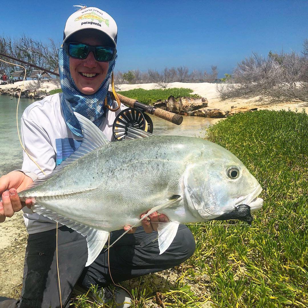 A person in a sun hat, sunglasses, and neck gaiter kneels by a beach, holding a large silver fish caught on a fly rod.