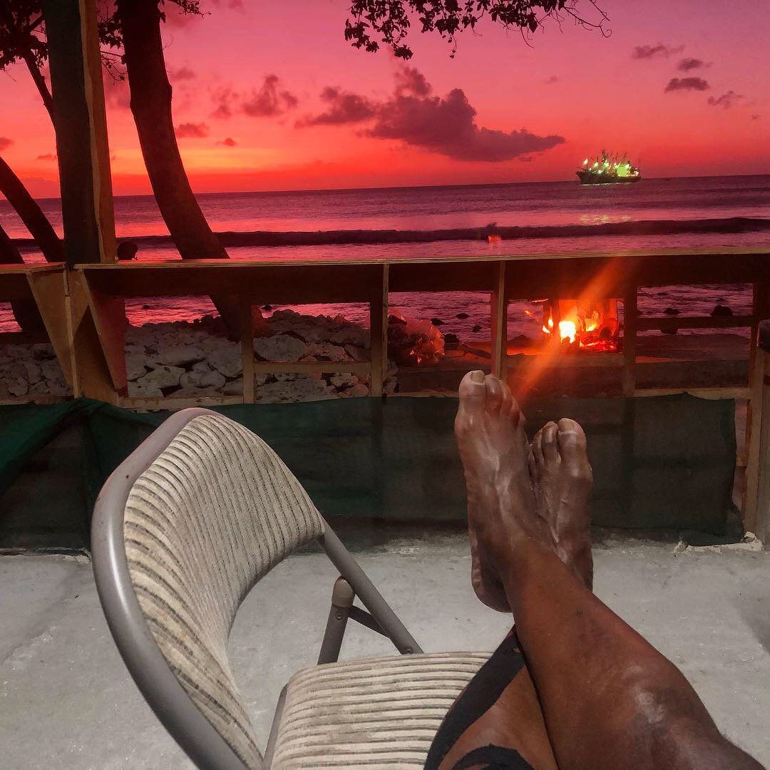 Bare feet resting on a chair overlooking a vibrant pink sunset over the ocean with a ship in the distance.