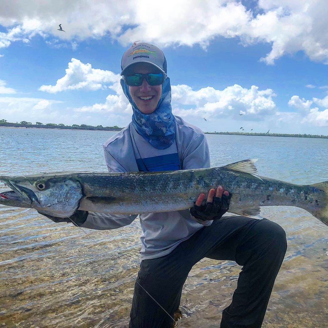 A person wearing a hat and sunglasses smiles while kneeling to hold a large barracuda near a body of water.