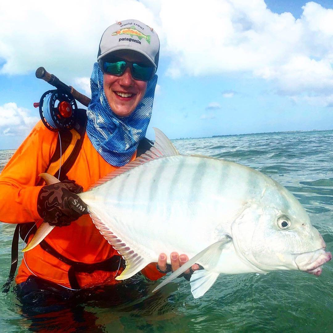 A smiling angler wearing a sun mask and hat holds a striped, silvery-white fish while standing in shallow ocean water.