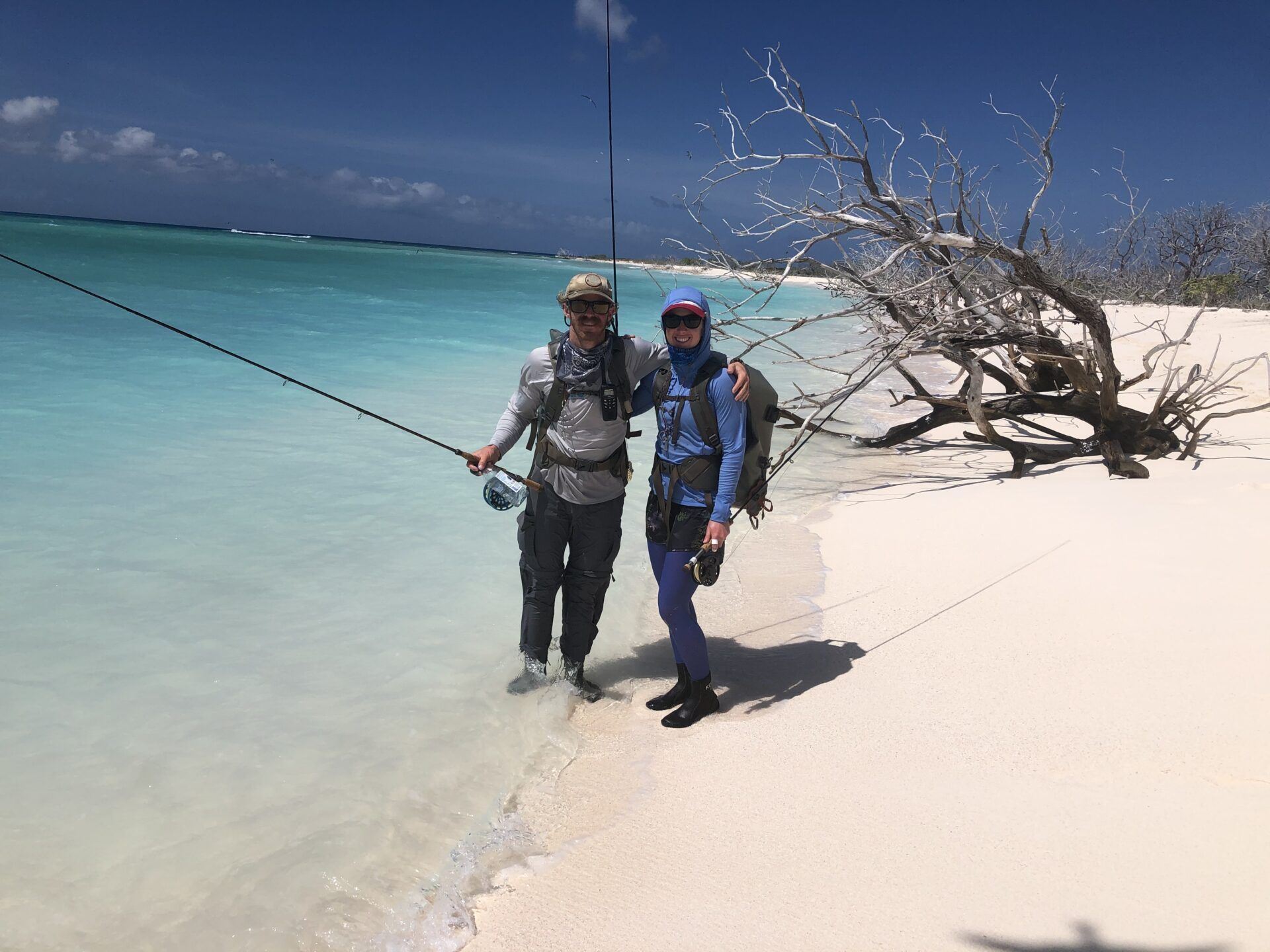 Two people in fishing gear stand on a bright white sandy beach next to a large piece of driftwood by turquoise water.