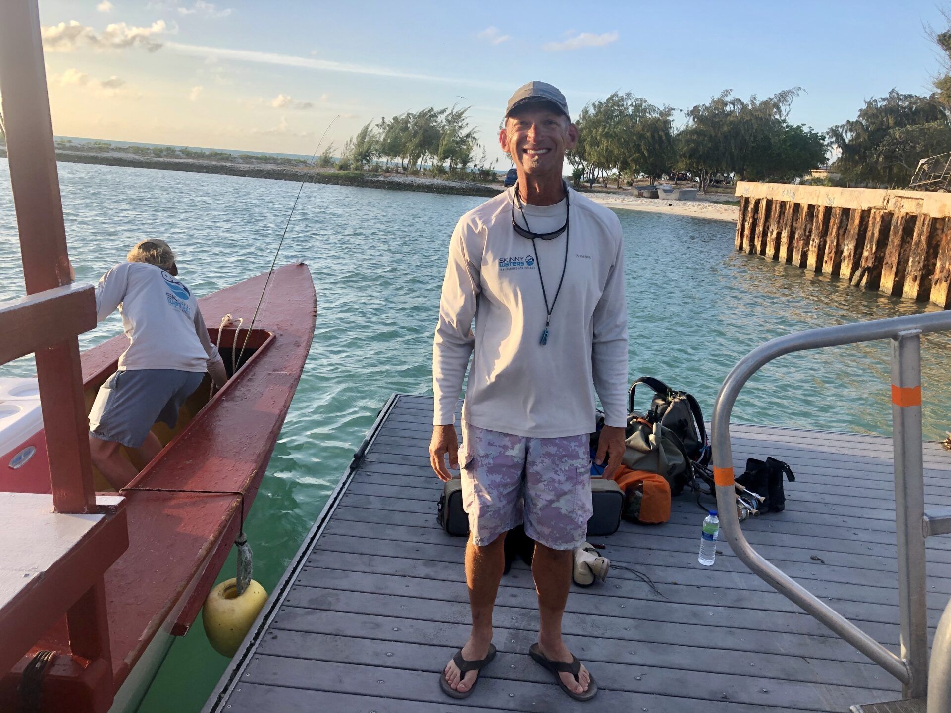 A smiling person in a long-sleeved shirt and shorts stands on a dock, with another person fishing from a boat nearby.