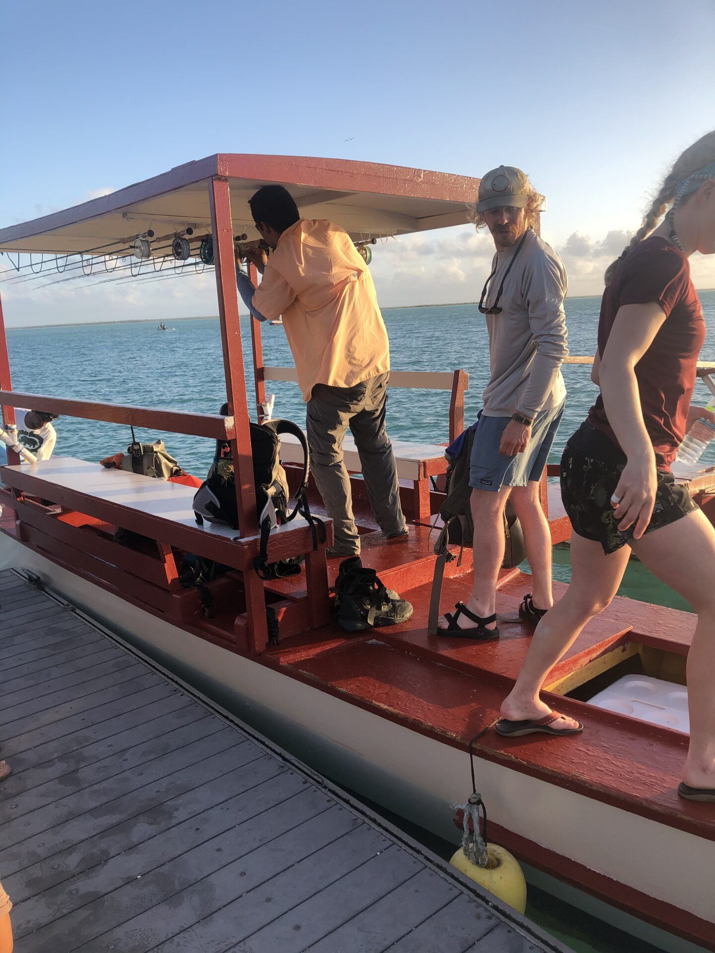 Three people board a red-roofed boat docked at a wooden pier on the water under a clear blue sky.