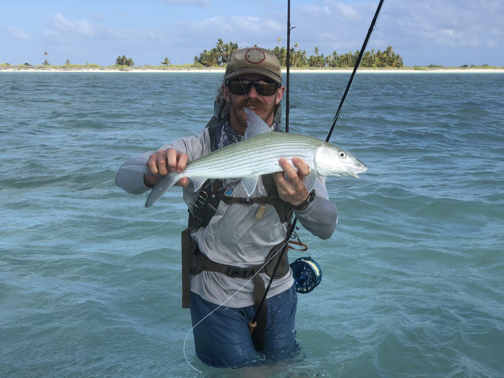A person wearing sunglasses and a hat stands in shallow turquoise water, holding a silver fish caught on a fly rod.