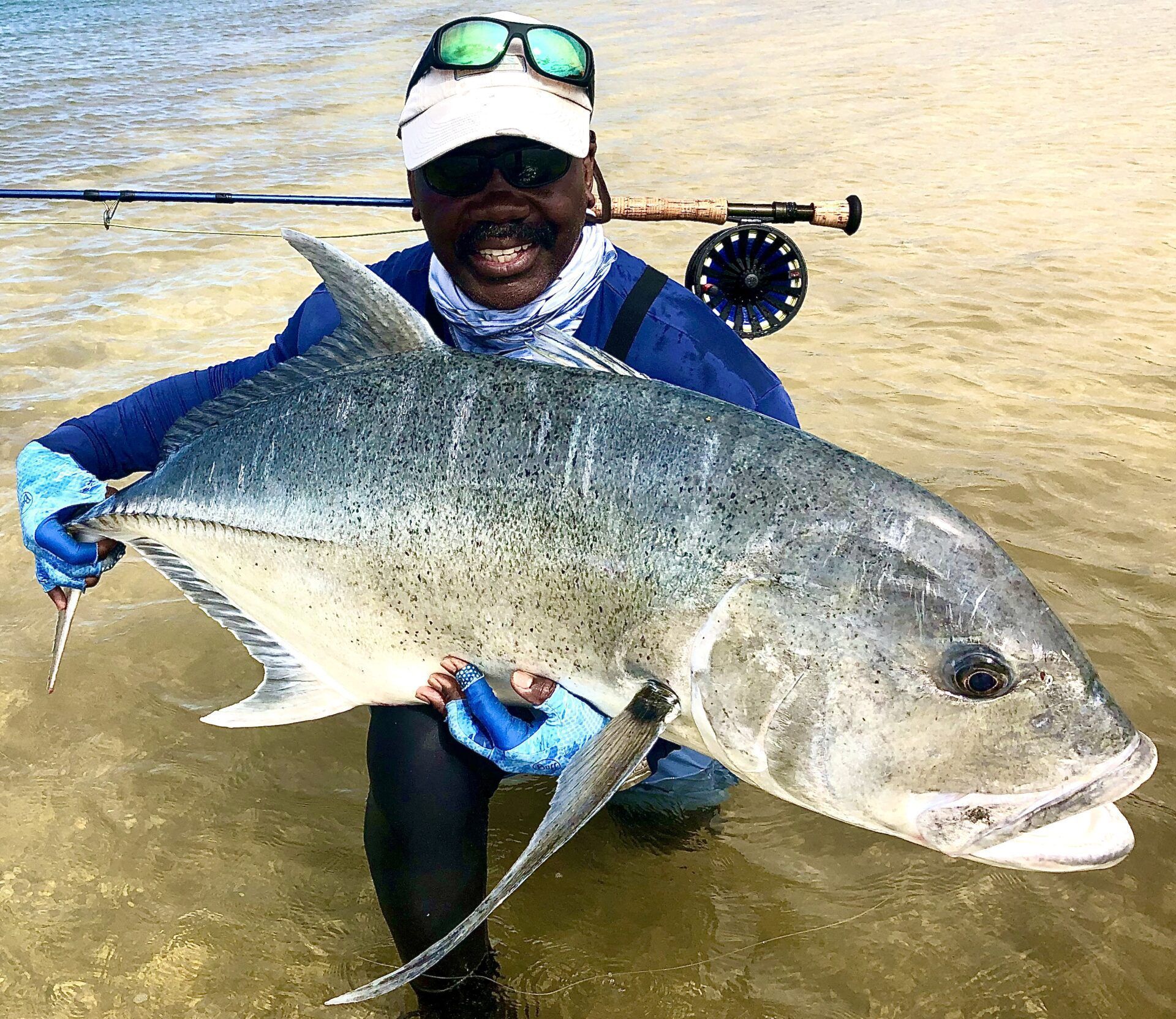 A person kneeling in shallow water, smiling while holding a large, grey trevally fish caught with a fly fishing rod.