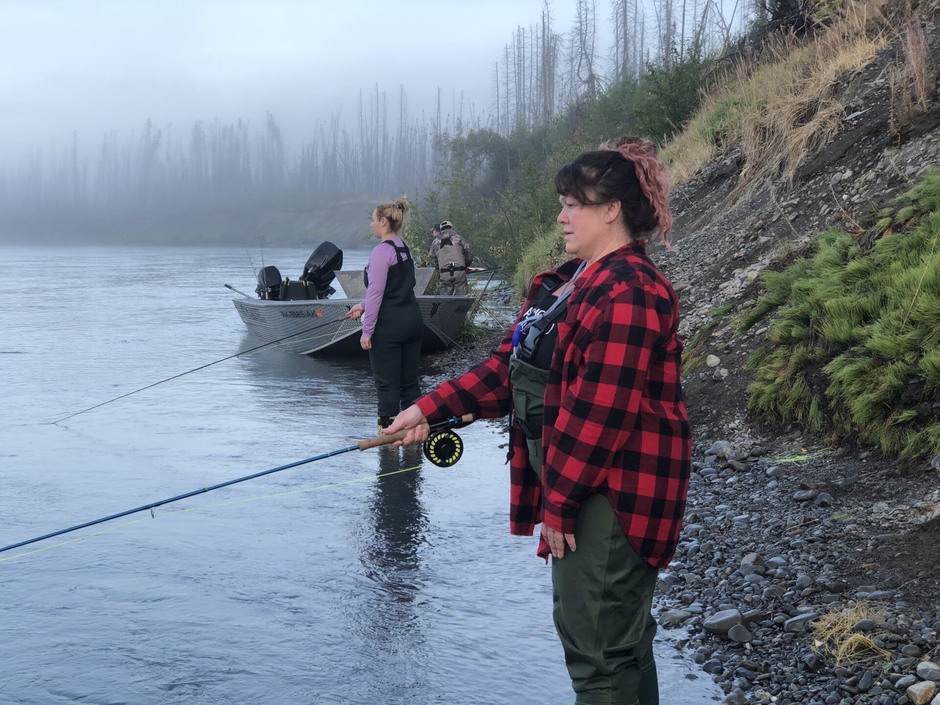 Two people in waders fish with rods from the rocky bank of a foggy river near a boat.