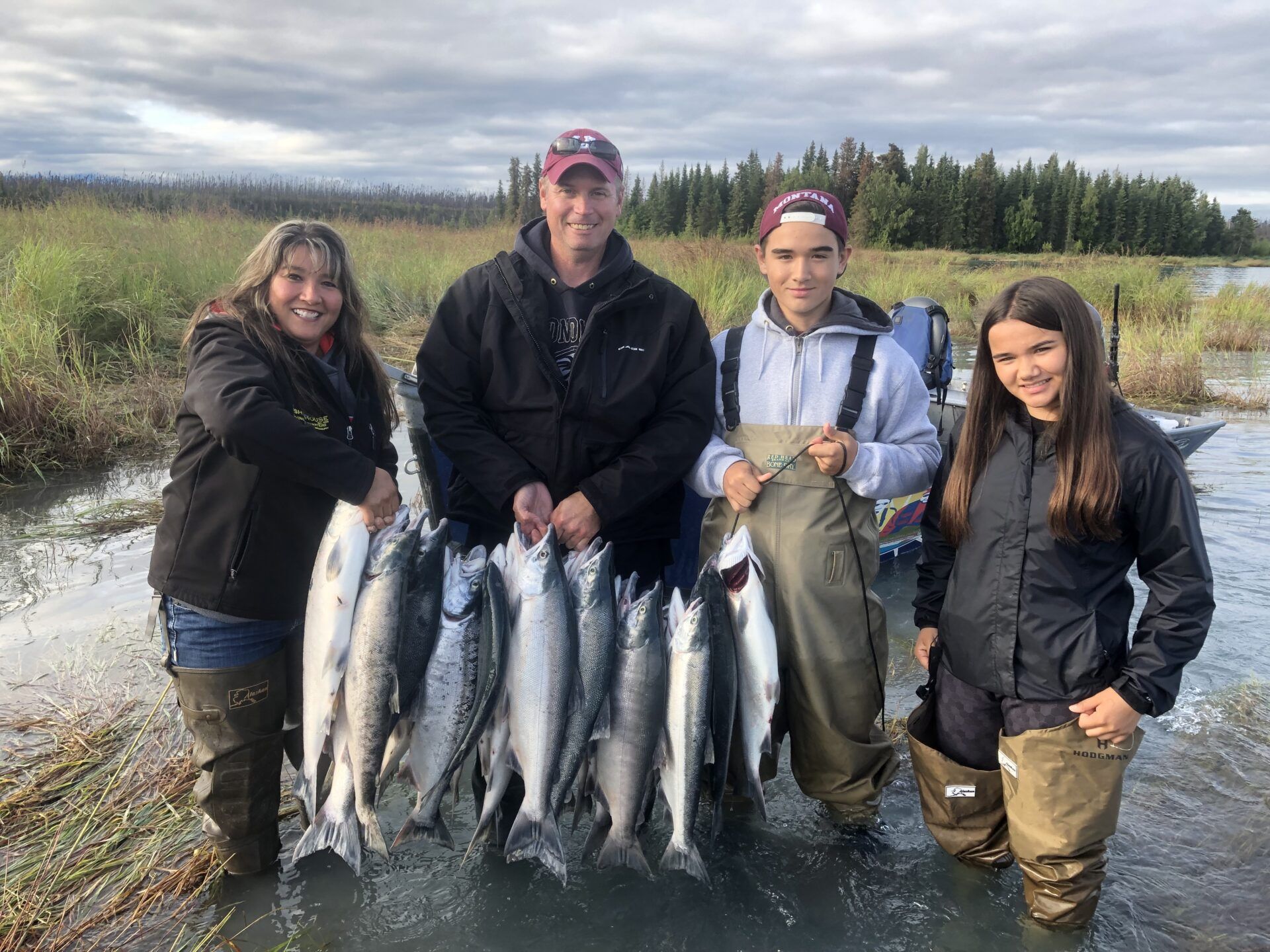 Four people in outdoor gear standing in water, holding several fish in a line, with a forest and cloudy sky in background.