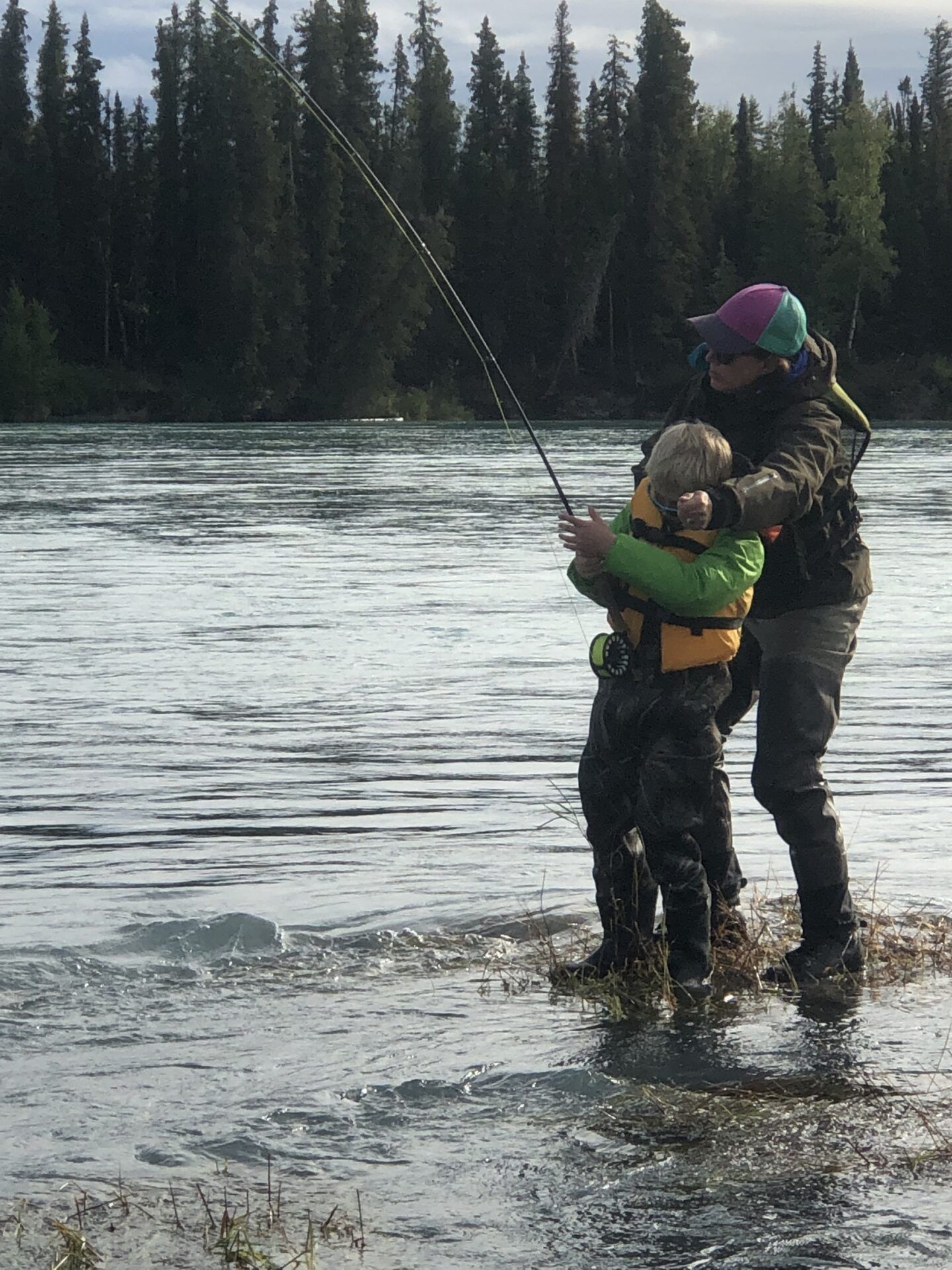 An adult and child in life vests fly-fish together while standing in a flowing river backed by a dense forest.