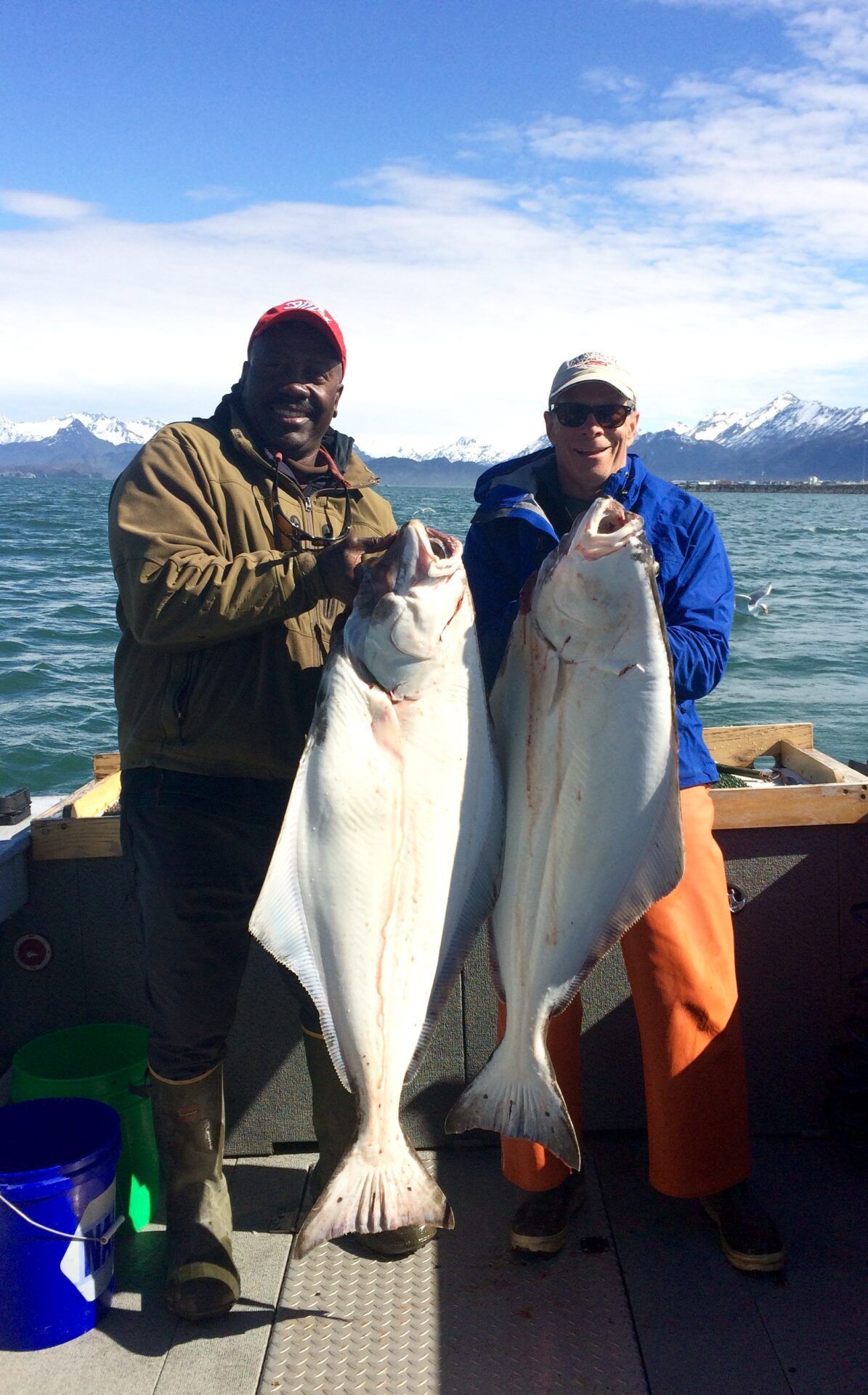 Two people stand on a boat deck, each holding a large halibut against a backdrop of snow-capped mountains and water.