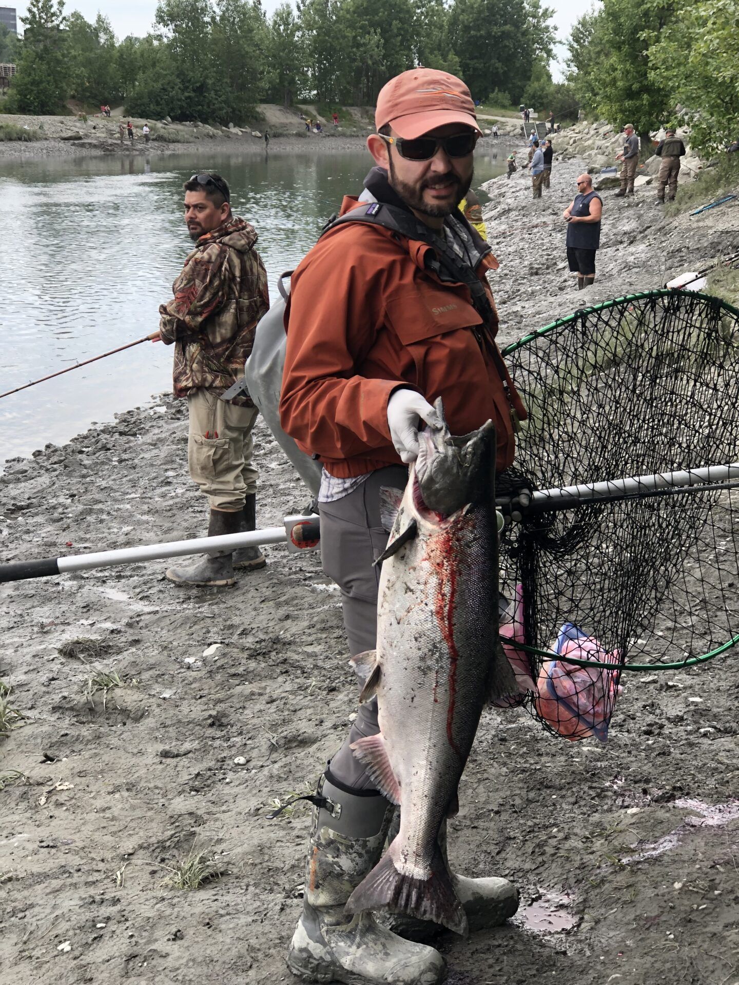 A person wearing a hat and orange jacket holds a large fish on a rocky riverbank, with other anglers nearby.