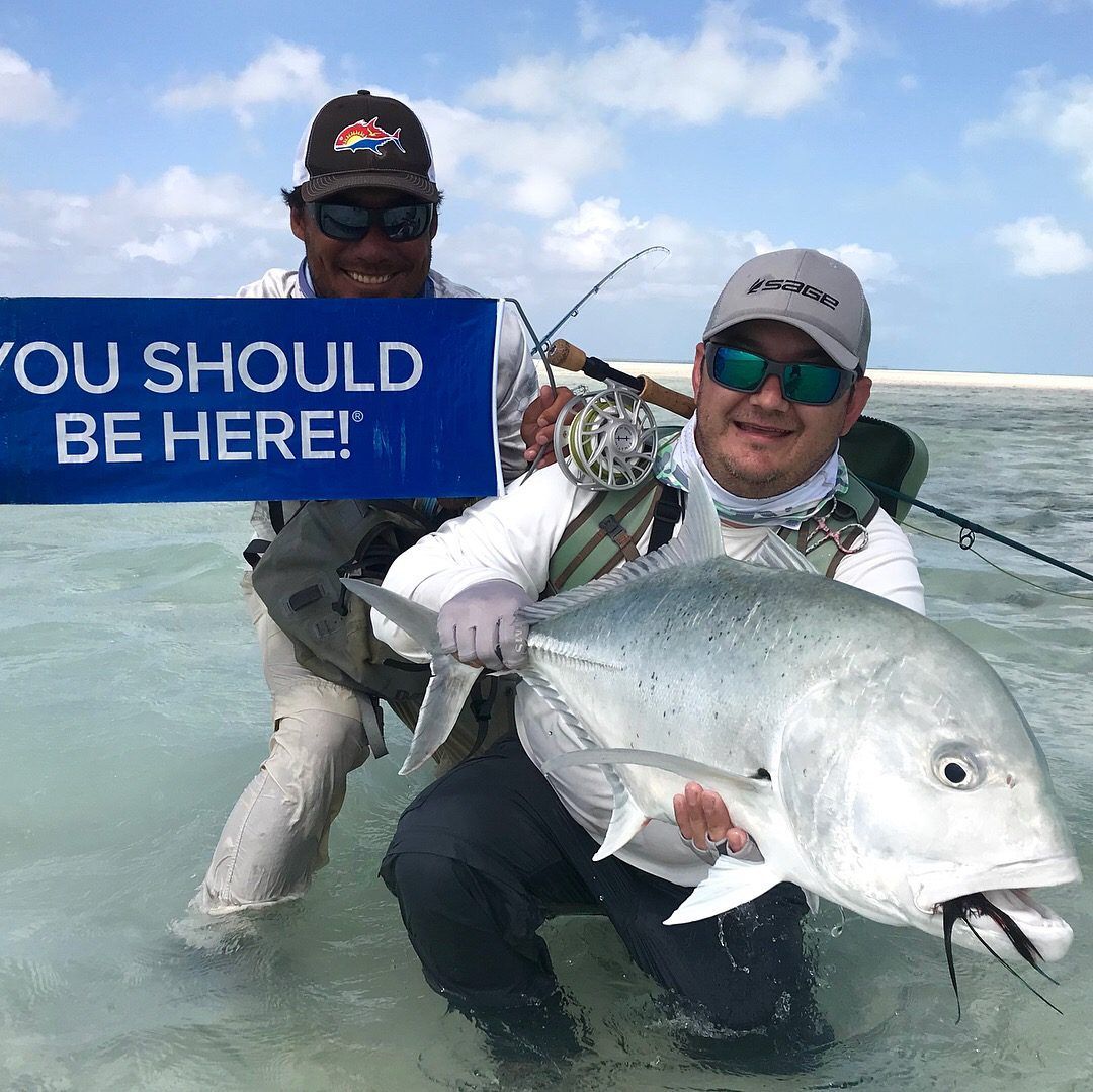 Two people in a shallow, clear ocean hold a large fish and a blue banner that reads 