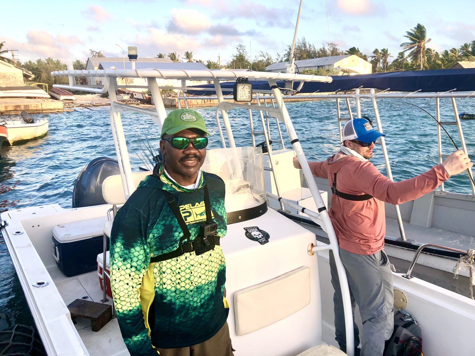 Two people on a boat in a harbor; one stands at the helm while the other stands behind holding a fishing rod.