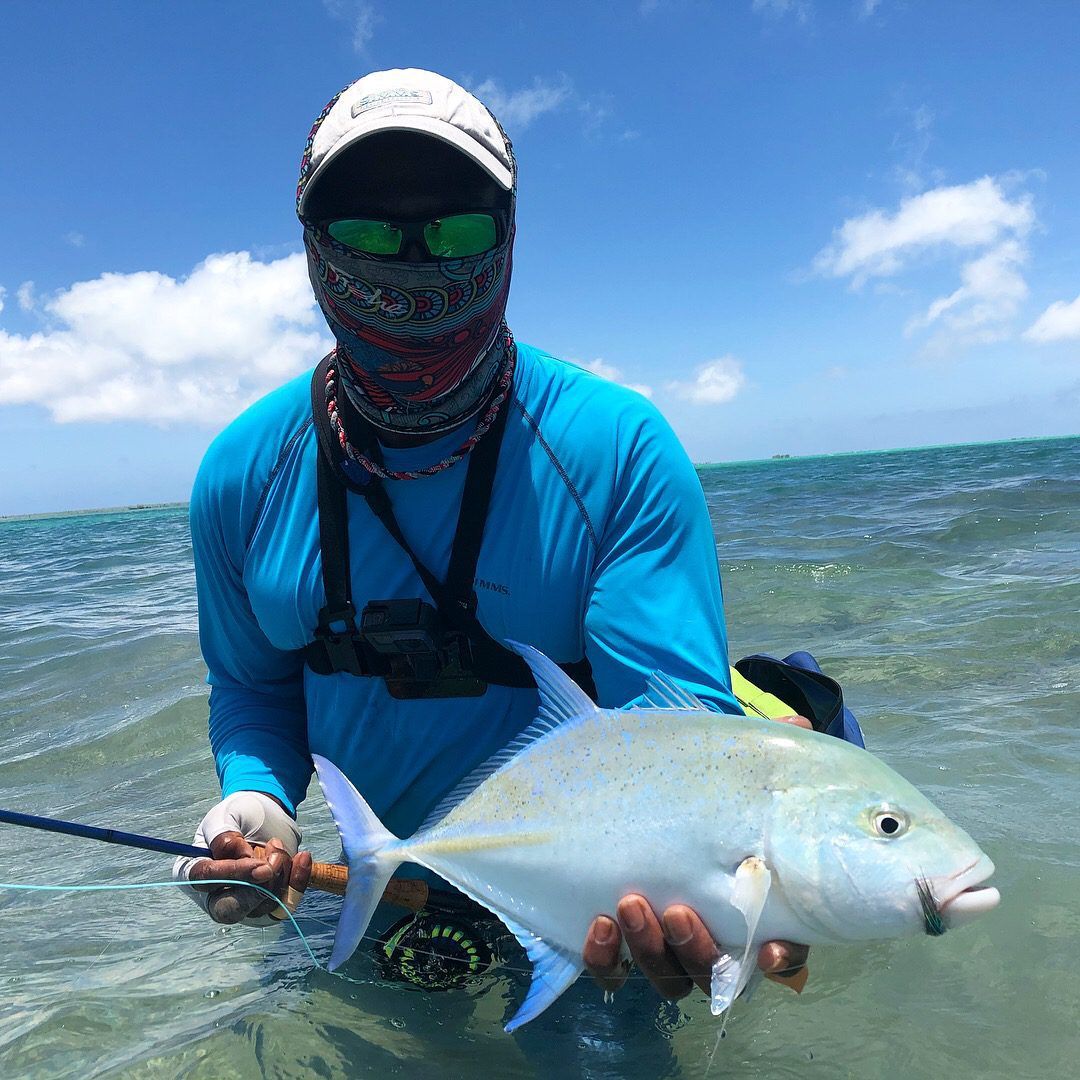 A person wearing protective gear stands in shallow water, holding a silvery fish they have caught.