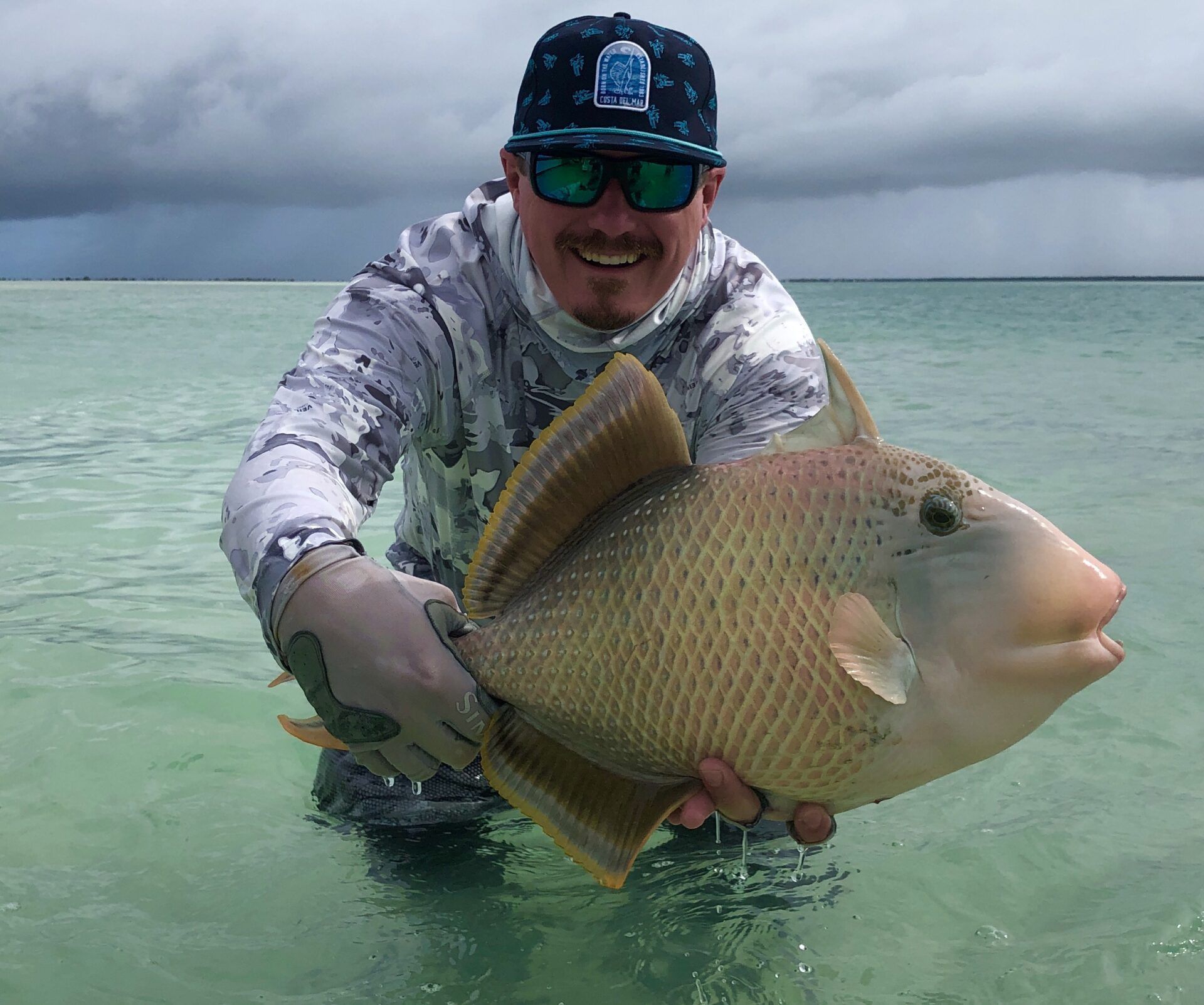 A person wearing a camo long-sleeve shirt and sunglasses holds a large, tan-colored triggerfish while wading in clear water.