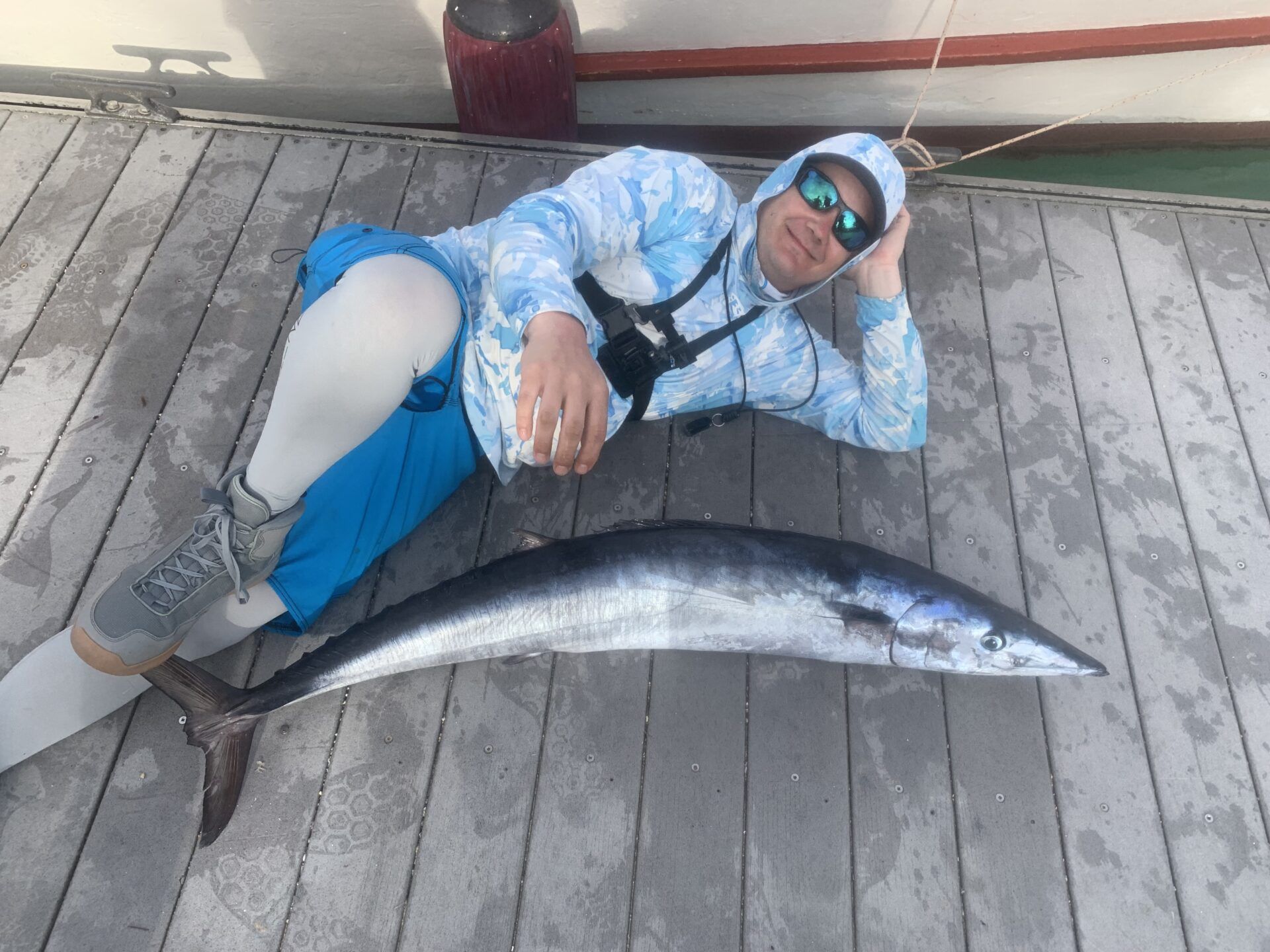A person wearing a light blue patterned hoodie, blue shorts, and sunglasses reclines on a wooden dock next to a large fish.