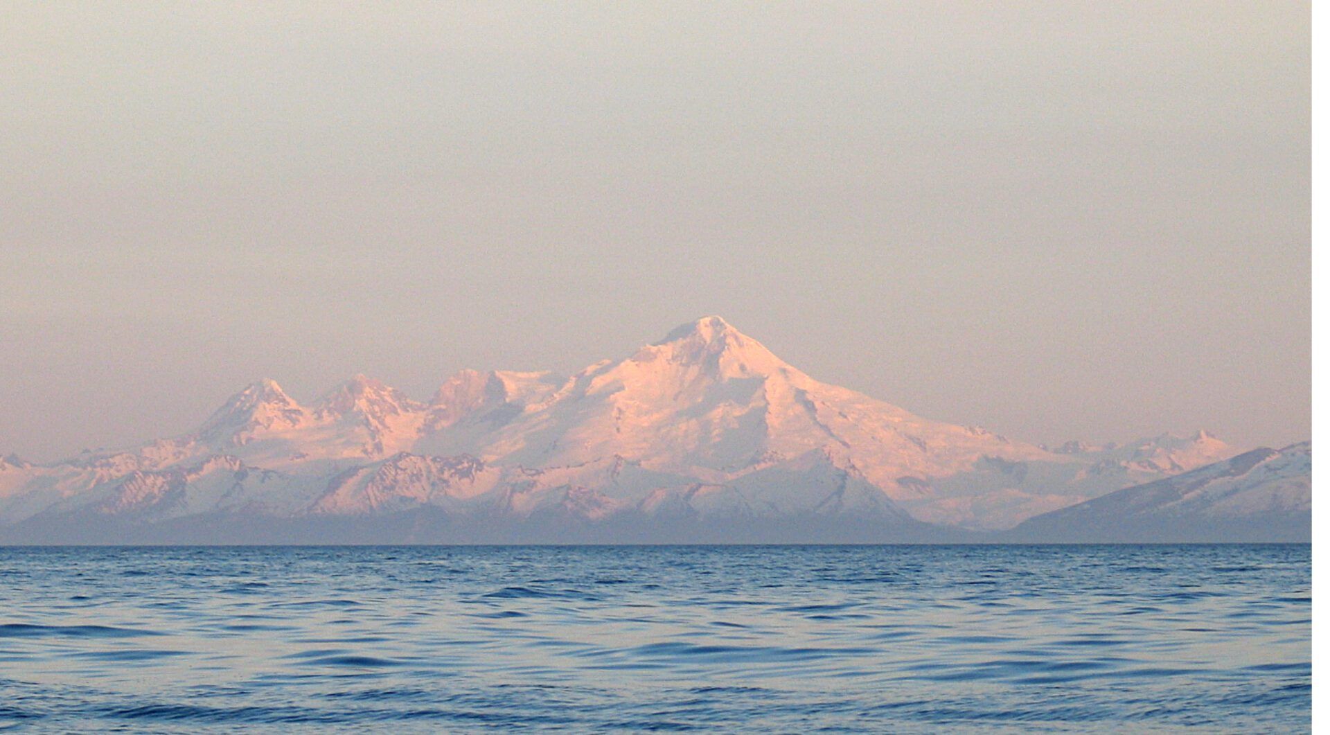A snow-capped mountain range rises behind calm, dark blue ocean waters at sunrise, bathed in a soft pink light.