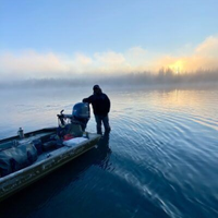 A person stands waist-deep in calm water beside a small aluminum boat at sunrise, surrounded by low-hanging fog.