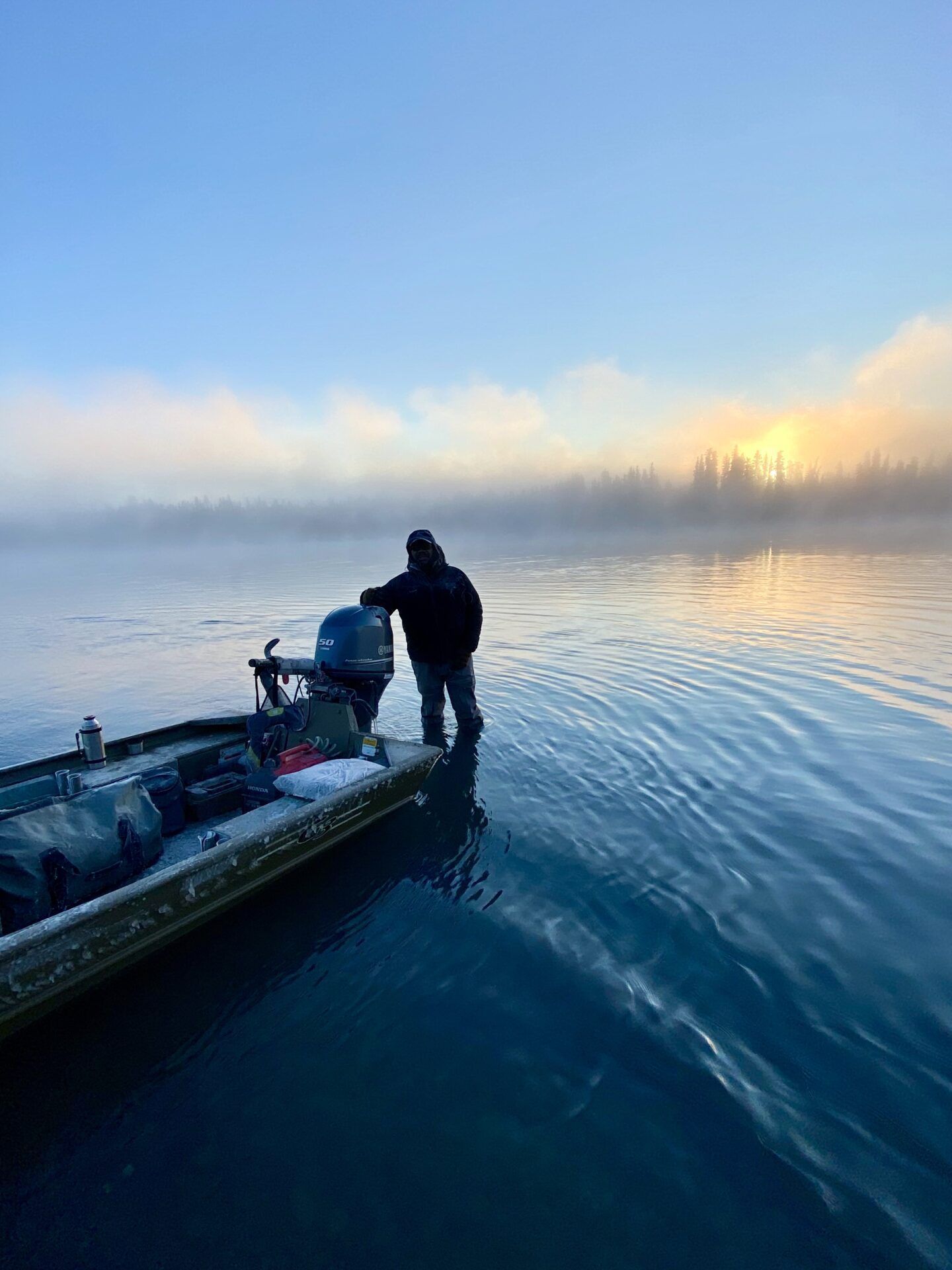 A person stands in shallow water next to a camo-patterned motorboat at sunrise, with light fog over the calm lake.