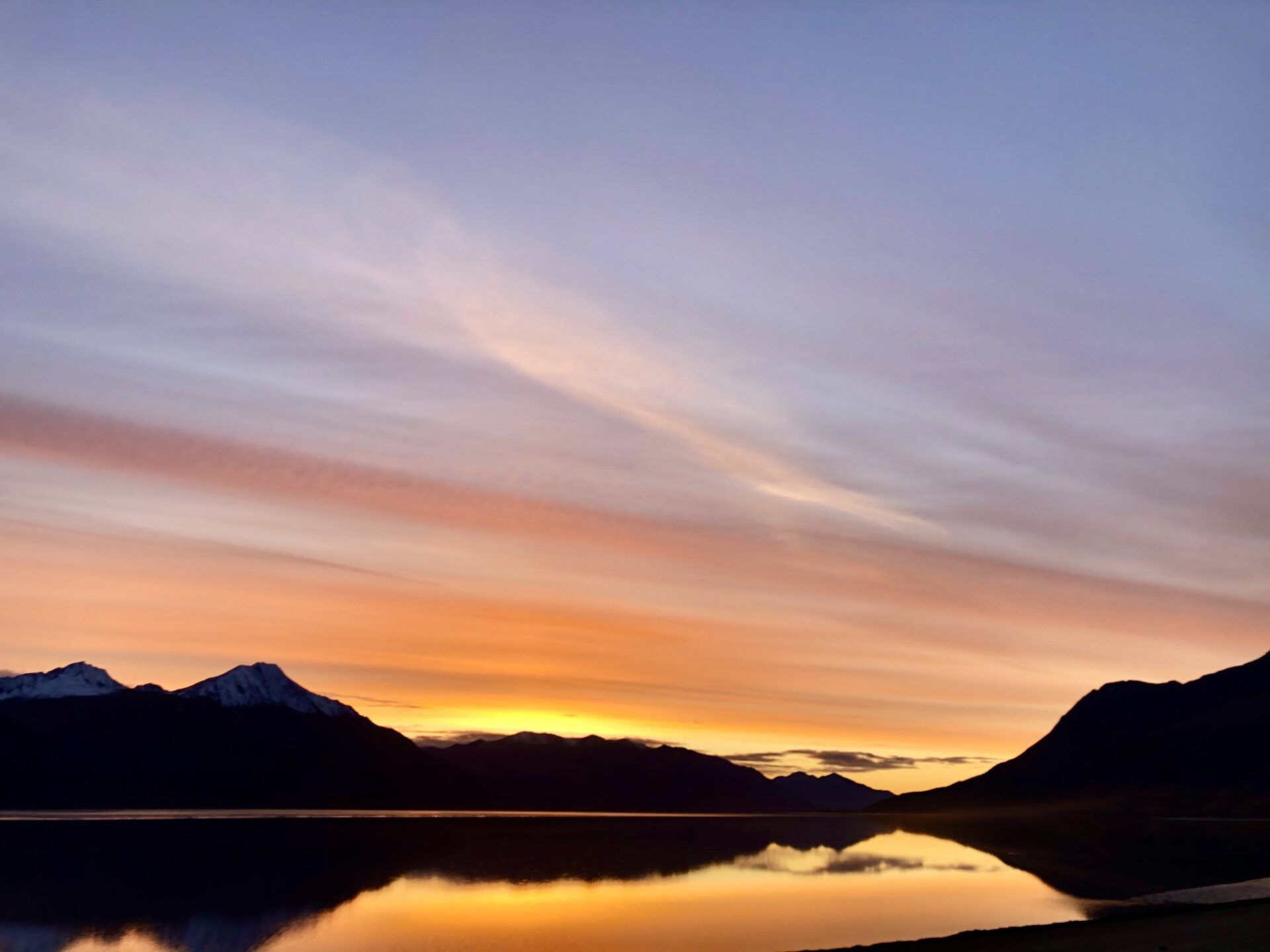 Sunset over a calm lake, with silhouetted mountains reflecting in the water under a soft, orange and purple gradient sky.
