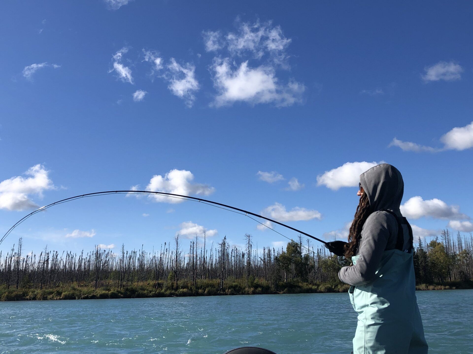 A person in waders and a hooded sweatshirt holds a fishing rod arched deeply over a river with trees in the background.