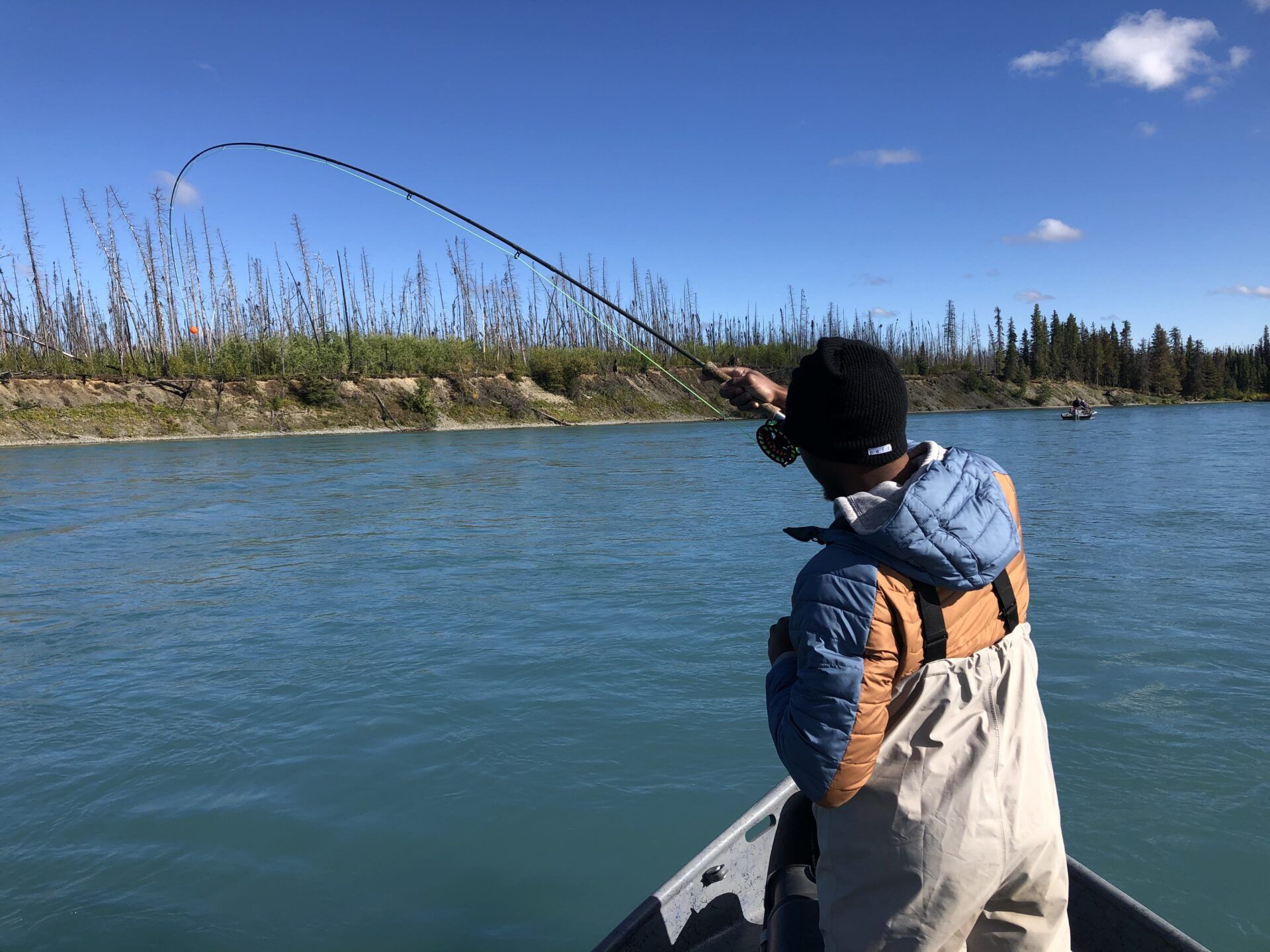 A person in waders and a jacket fly fishing from a boat on a river, with their rod arched over the water.