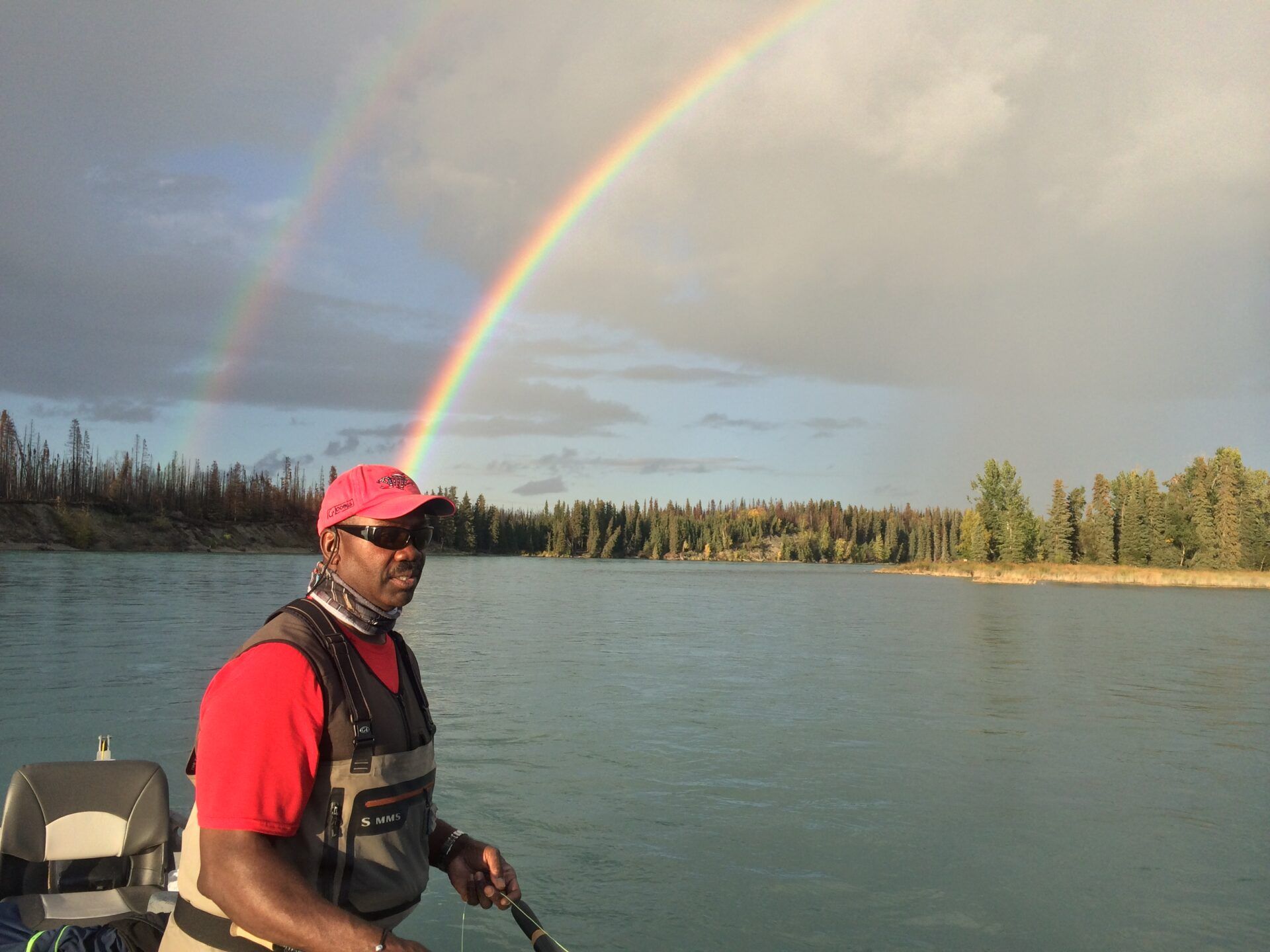 A person wearing a hat and fishing vest on a boat on a river, with a double rainbow arching across the cloudy sky.