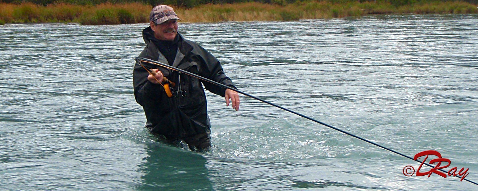 A person in a dark waterproof jacket and cap stands in a blue river, holding a fishing rod while fly fishing.