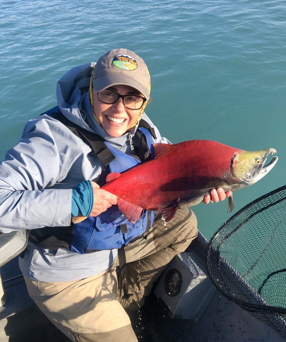 A smiling person in a blue life vest holding a large, vibrant red salmon in a boat on blue water.