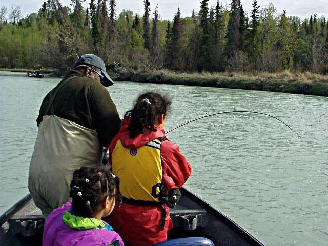 An adult and two children on a boat in a river, with the older child holding a fishing rod bent by a catch.