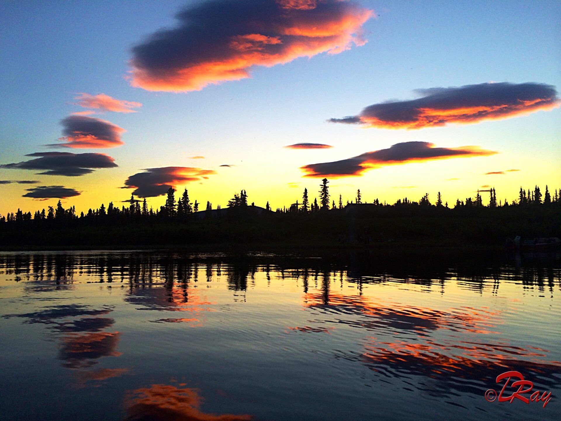 A sunset glows over a calm lake, with dark tree silhouettes reflecting on the water and orange-tinted clouds above.