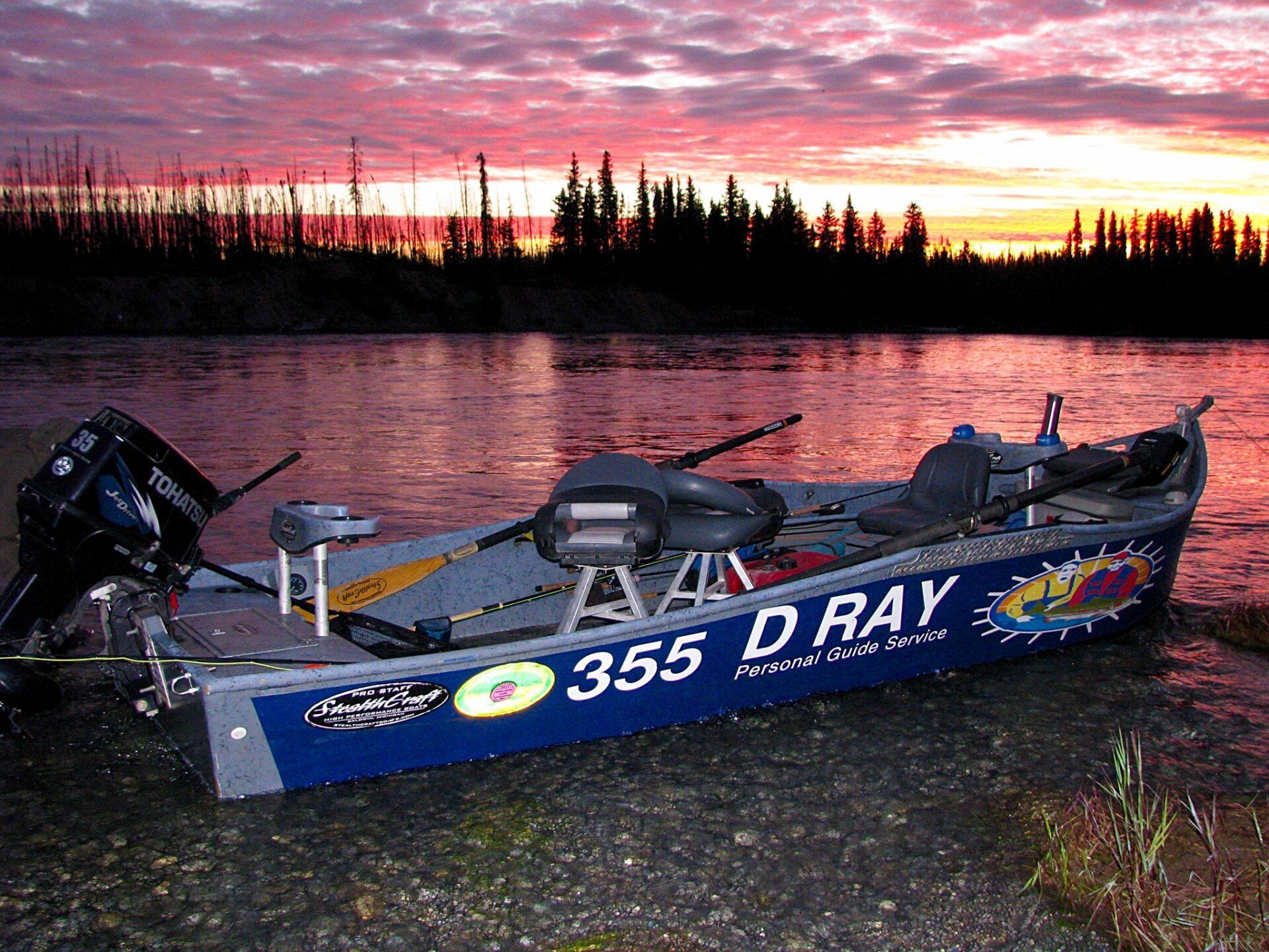 A blue motorized fishing boat rests on a rocky shore at dusk with a vibrant pink and orange sunset over the water.