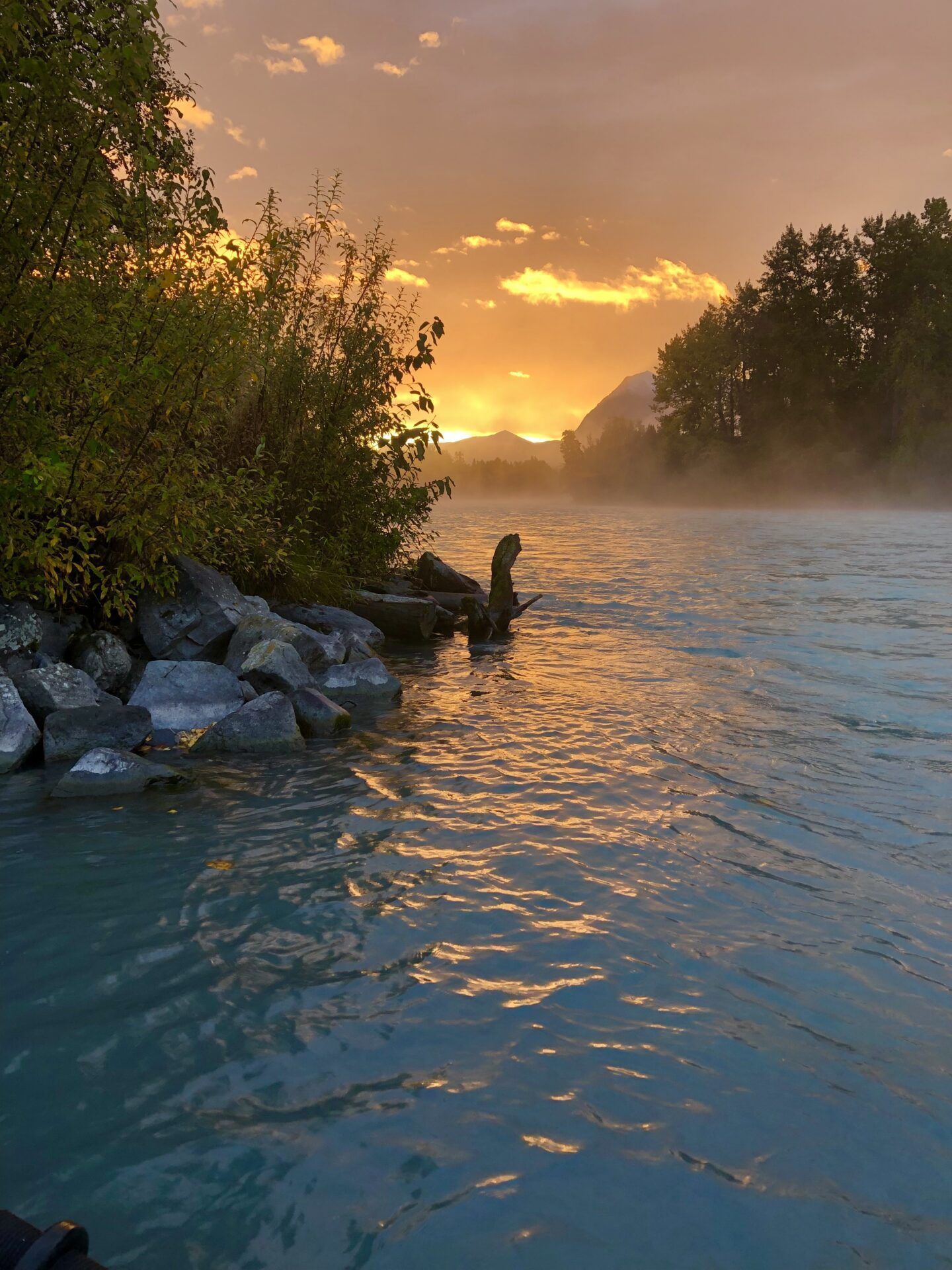 A river flows toward a mountain range under a golden, hazy sunset, with rocky banks and trees in the foreground.