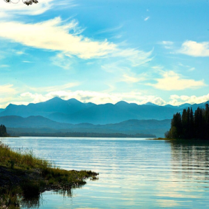 A calm lake reflects a bright blue sky, bordered by a dark forest shoreline with majestic, hazy mountain peaks beyond.